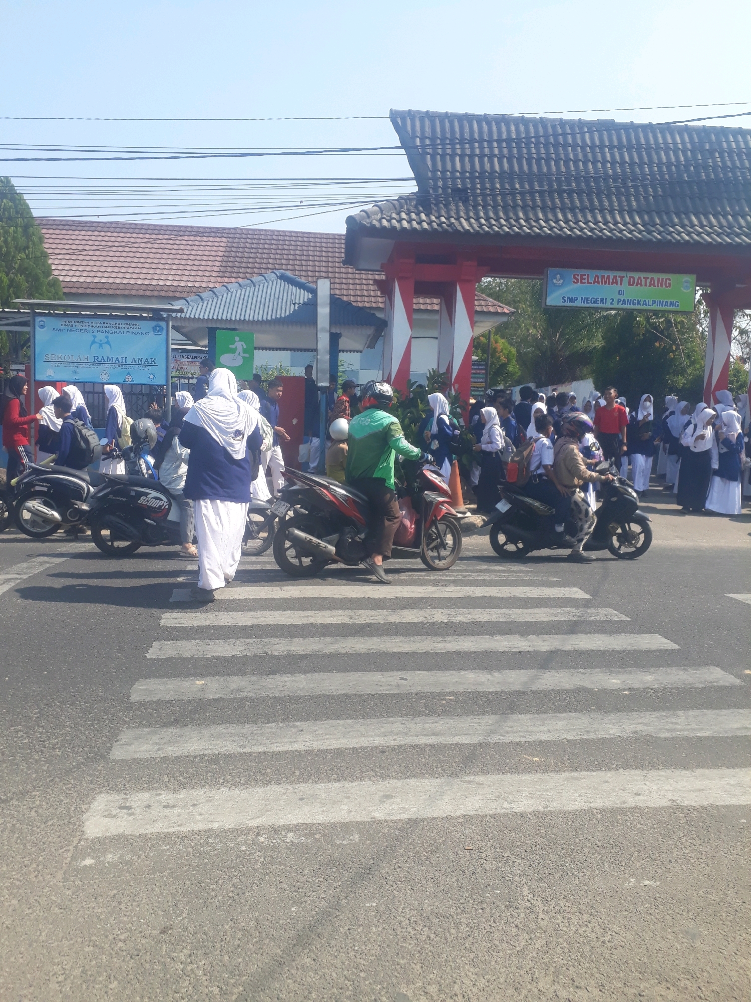 The image depicts a busy scene outside SMP Negeri 2 Pangkalpinang, located in the city of Pangkalpinang, Indonesia. A pedestrian crossing is prominently visible in the foreground, with several motorcycles and scooters on or near it. A group of students, many wearing hijabs, are gathered on the sidewalk and in the street, some appearing to be waiting to cross or having just disembarked from vehicles. One individual in a green jacket and helmet is on a scooter in the middle of the crossing, while another person in a brown jacket and patterned pants is also on a scooter, carrying a passenger. A large banner with the text "SEKOLAH RAMAH ANAK" (Child-Friendly School) is visible on the left, and another banner that reads "SELAMAT DATANG DI SMP NEGERI 2 PANGKALPINANG" (Welcome to SMP Negeri 2 Pangkalpinang) is displayed above an archway. The architecture suggests a school entrance, with a tiled roof and red and white striped pillars. The environment appears to be urban, with overhead wires and trees visible in the background, under a clear blue sky.