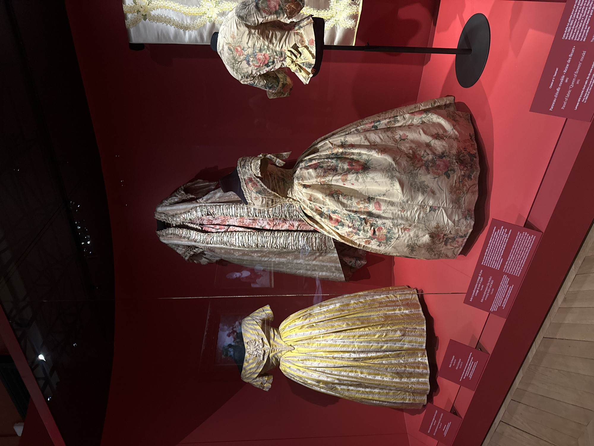 The image shows a display of historical clothing at an exhibition, potentially in Paris, France. On a red background, three dresses or dress components are showcased. The top right consists of a bodice mounted on a dark rod, with floral print on light base. The central dress, also of a light shade with a similar floral pattern, is more complete, showing a fuller skirt. The bottom dress is yellow with narrow vertical stripes and a smaller bow detail. Alongside each piece are informational plaques in French. The floor is wooden.