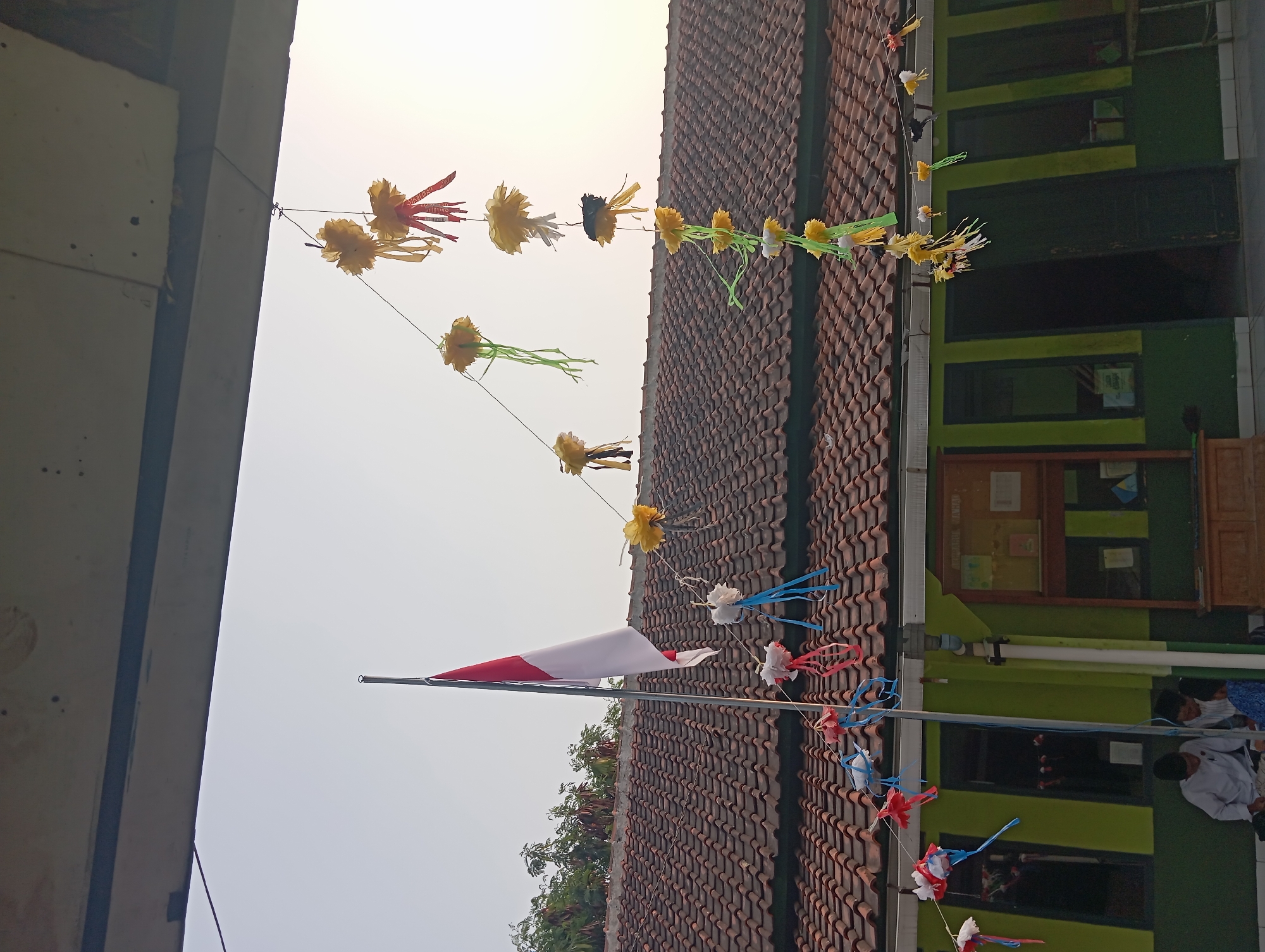 A low-angle, medium shot captures the exterior of a single-story building with a dark, tiled roof. The building is painted green with lighter green accents around the windows and doors. A flagpole with the Indonesian flag, red and white, stands prominently to the left of the frame. Decorative streamers, made of yellow and white paper flowers with red and blue ribbon tails, are strung across the scene. The streamers hang from wires attached to an overhang above the building. In the foreground, three young males, two wearing white shirts and one in a blue patterned shirt, sit on the ground in front of the building. One boy is looking down at a book or papers, while the other two appear to be interacting. The sky is clear and pale blue, suggesting it is daytime. The overall impression is one of a school or community building in Nanjungmekar, Indonesia, perhaps decorated for a celebration or event.