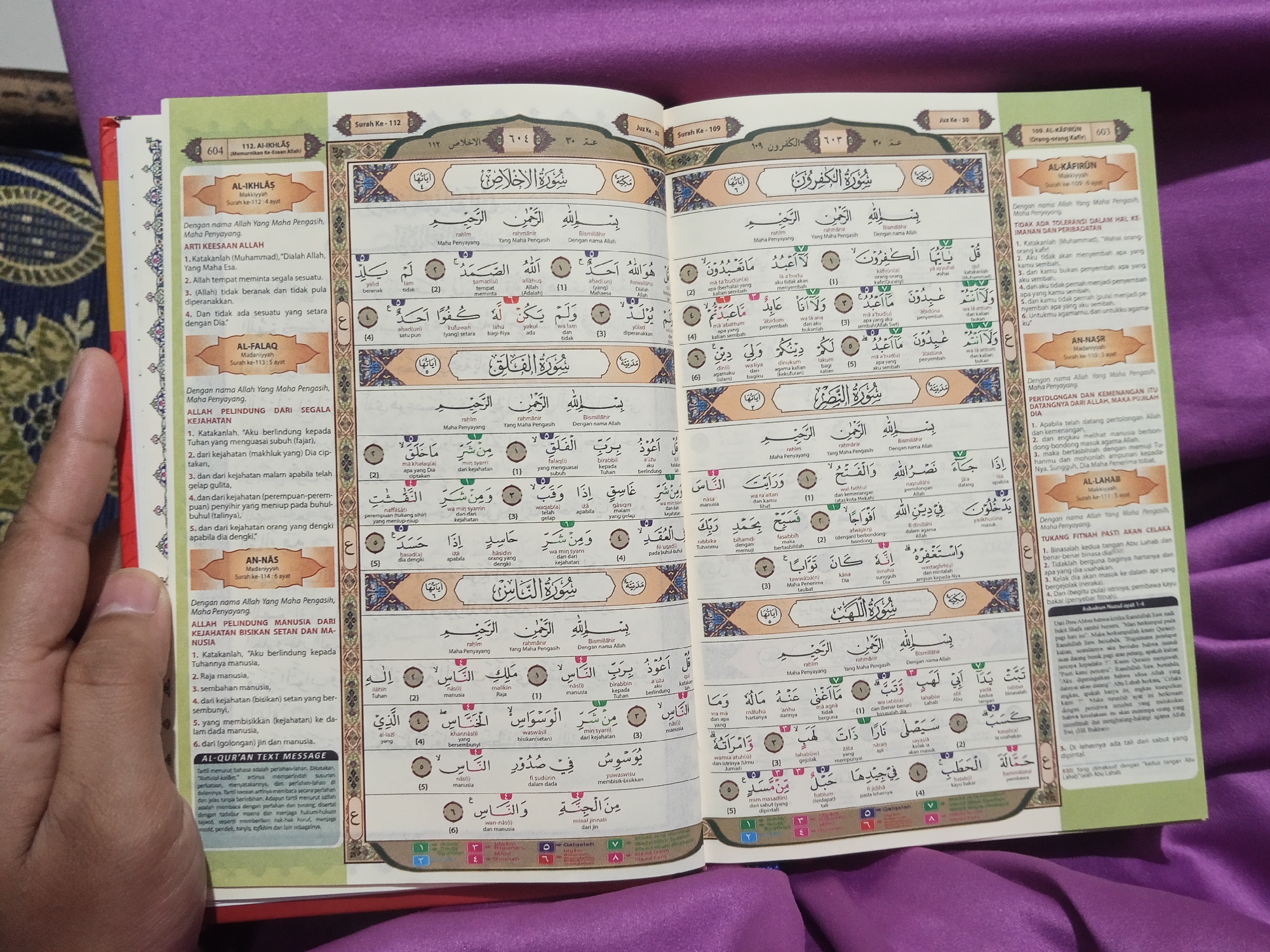 The image shows an open book, likely a Quran, displayed on a dark surface. The book is open to pages displaying Arabic script and translations. The left page features Surah Al-Ikhlas and Surah Al-Falaq, along with their meanings and verses. The right page displays Surah Al-Kafirun, Surah An-Nasr, and Surah Al-Lahab. The surrounding area is out of focus but appears to be a textile material. The book itself is the primary subject, with the text and layout being the main points of interest. There are no people or specific environmental details that would indicate a location in Lestari Indah, Indonesia.