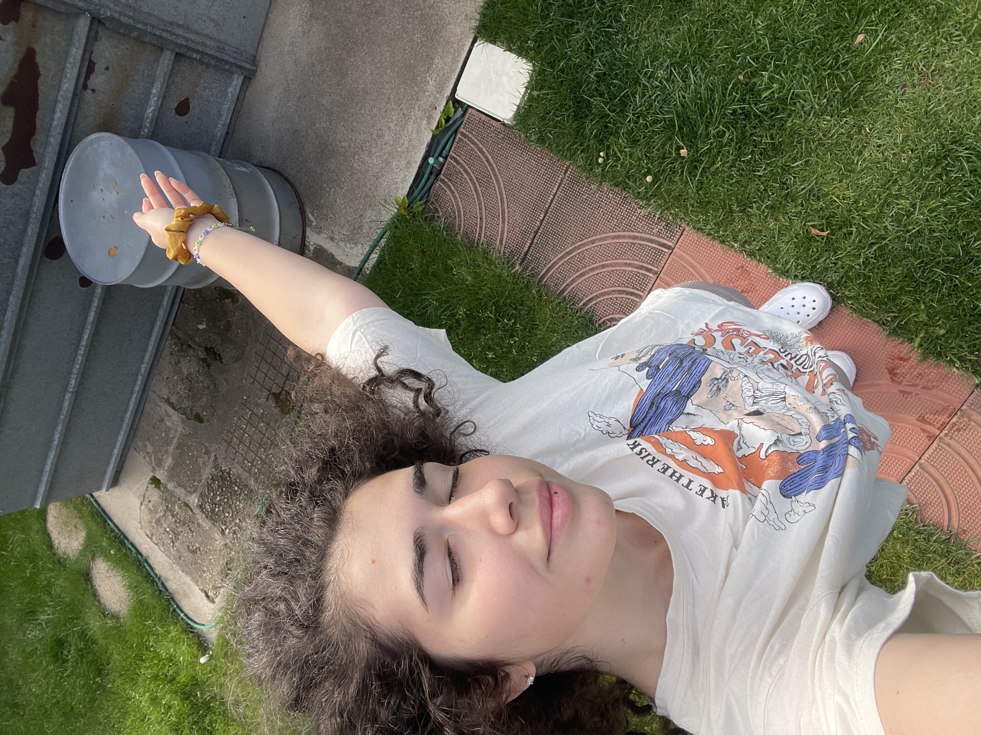 The image captures a high-angle, portrait-oriented view of a young person lying relaxed in an outdoor setting, likely a garden or backyard. The subject has curly dark brown hair and fair skin with visible freckles or moles. Her eyes are closed, and she wears a calm, serene expression, featuring a small stud piercing on her philtrum.
She is dressed in a white t-shirt adorned with a colorful graphic design in blue, orange, and white, depicting stylized figures or scenes with clouds. The phrase "TAKE THE RISK" is clearly readable on her shirt. On her left wrist, she wears a yellow scrunchie and a delicate beaded bracelet. Her feet, clad in white Crocs, are visible. Her left arm is extended upwards and to the left, pointing towards a large, cylindrical grey metal barrel that shows signs of rust.
The ground features a mix of lush green grass, sections of concrete, and reddish-brown patterned stepping stones or mats. A green garden hose is visible along the edge of the grass and concrete. The bright, natural light suggests a sunny day, possibly morning or late afternoon, creating a peaceful ambiance. The scene is set in an outdoor private space, consistent with a garden in Sofia, Bulgaria.