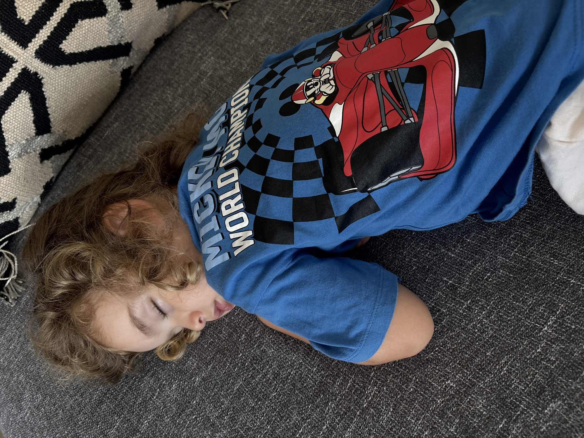 A young child with curly blonde hair is sleeping on a gray couch, their face turned away from the camera. They are wearing a blue t-shirt with a graphic of Mickey Mouse in a race car and the text "MICKEY WORLD CHAMPION" visible on the shirt. The child's eyes are closed, and their mouth is slightly open. The couch has a textured, woven fabric. To the left of the child, a portion of a throw pillow with a black and white geometric pattern is visible. The lighting suggests it could be daytime, but no specific weather cues are present. The scene appears to be indoors, likely a living room. There are no obvious indicators of the specific location within Dubai.