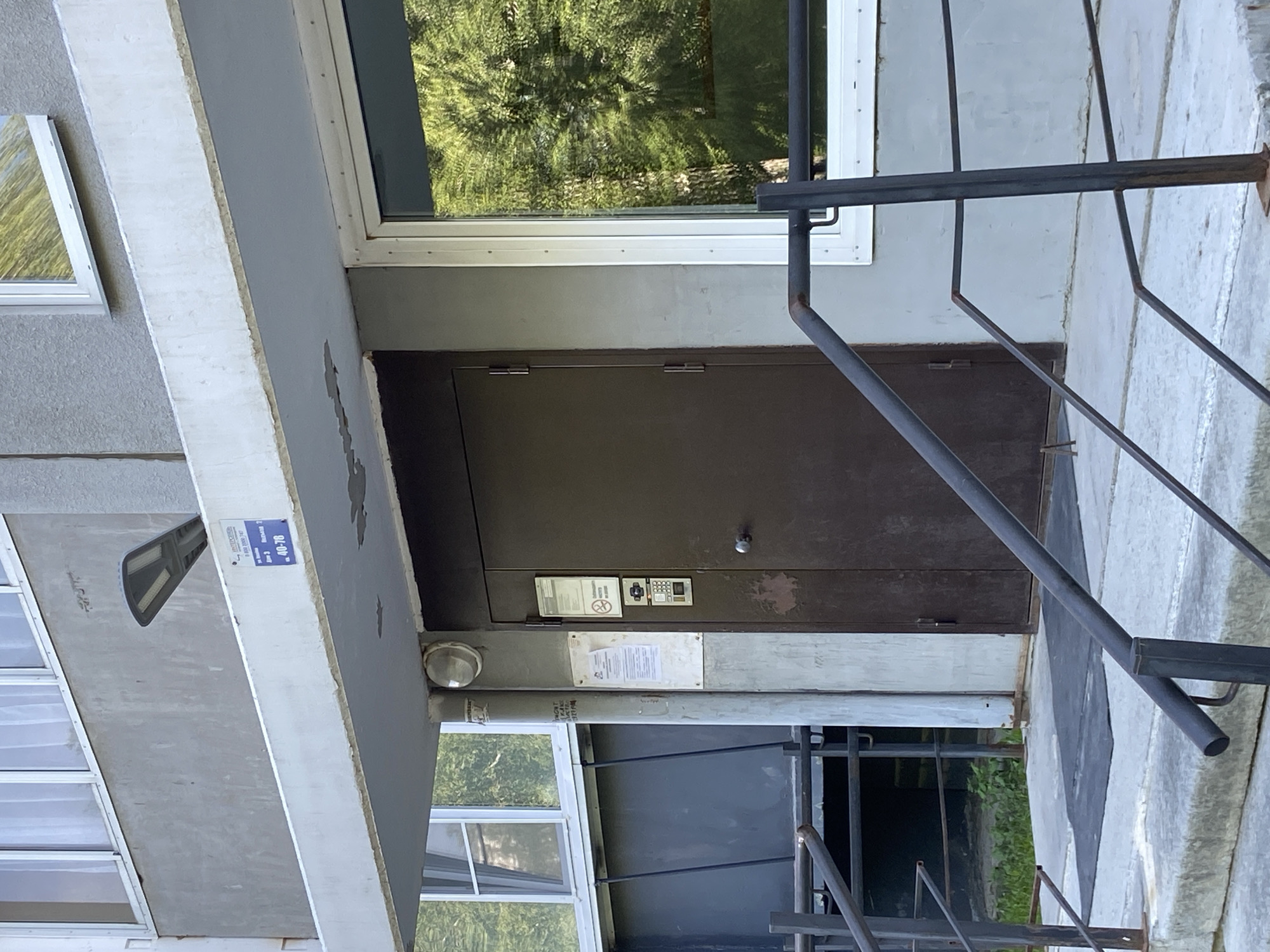 The image shows the entrance to an apartment building. Above the entrance is a balcony with a sign that reads "40-76". There is a modern-looking LED light fixture mounted on the underside of the balcony. A metal railing leads up to the entrance, which has a dark brown metal door with a doorknob and a keypad entry system. To the left of the door, there are two posters attached to the wall. The building's facade is made of gray concrete, and there is a window to the right of the door reflecting green trees and sky. The location is in the city of Chelyabinsk, Russia.