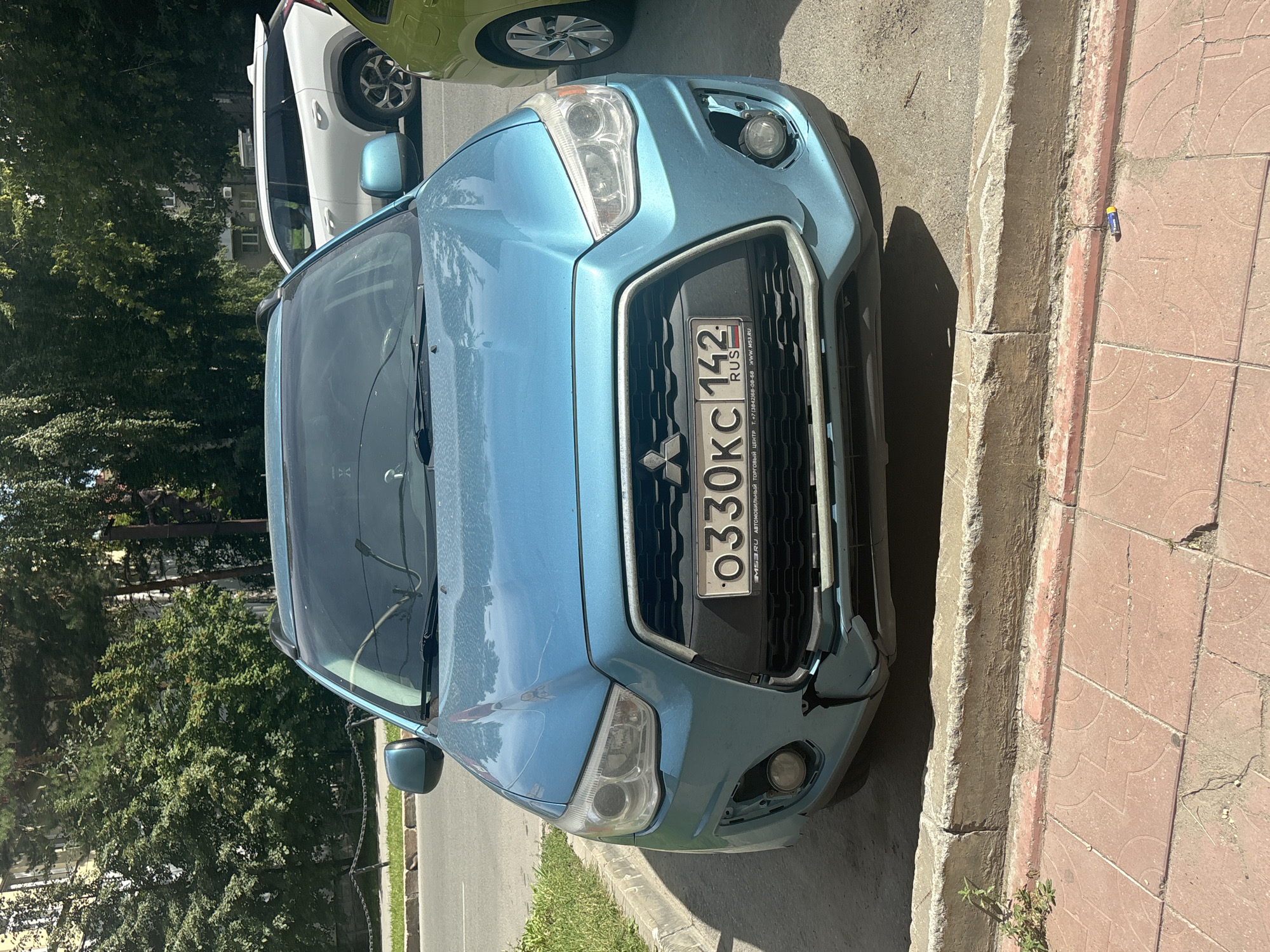 A blue Mitsubishi Outlander SUV is parked on a city street in Novosibirsk, Russia. The car is facing the camera, and its front bumper is damaged. The license plate reads "0330KC 142". A white SUV and a green car are parked behind it. The background shows trees and buildings. The street is paved with asphalt and bordered by a sidewalk made of red tiles and a concrete curb. There is some patchy grass and a small plant growing near the curb. The lighting suggests it is daytime.
