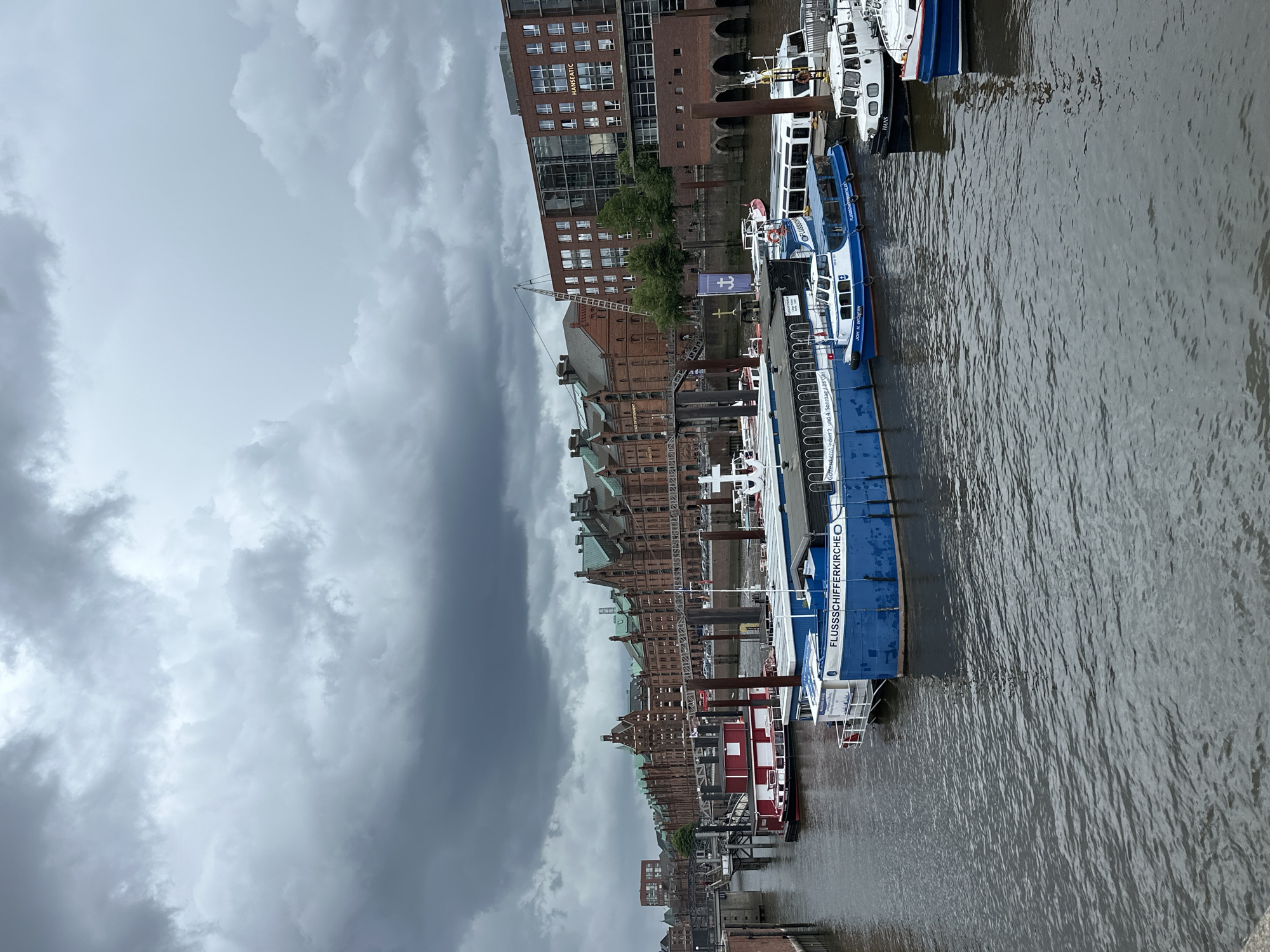 This image captures a bustling waterfront scene in Hamburg, Germany, under an overcast sky. The sky is filled with heavy, dark gray clouds, suggesting a cloudy day.
The foreground features dark, rippled water, indicative of a river or canal. Several boats are docked along the bank. The most prominent is a large, blue and white boat with "FLUSSSCHIFFERKIRCHE" (River Boat Church) written along its side. A sign on this boat also reads "Gottesdienst jeder 2. und 4. Sonntag / 11 Uhr" (Service every 2nd and 4th Sunday / 11 AM), indicating its function as a floating church. Another name visible on this boat is "JOH. H. WICHERN". To the left of the blue boat, a smaller red and white boat is docked. Further along, another white boat has "HANS" visible on its hull.
On the left side of the image, the riverbank is lined with historic brick buildings, characteristic of Hamburg's Speicherstadt district. These buildings, with their distinctive gabled roofs and numerous windows, reflect a blend of industrial and commercial architecture. Some have cranes, hinting at past or present port activities. A more modern brick building is also visible, with "HANSEATIC" written on its facade. Green trees dot the landscape between some of the buildings. A blue flag with a white anchor symbol is also visible near the waterfront.
The overall impression is that of a historic port city, with ongoing river traffic and a strong maritime heritage, enduring a typical cloudy day.