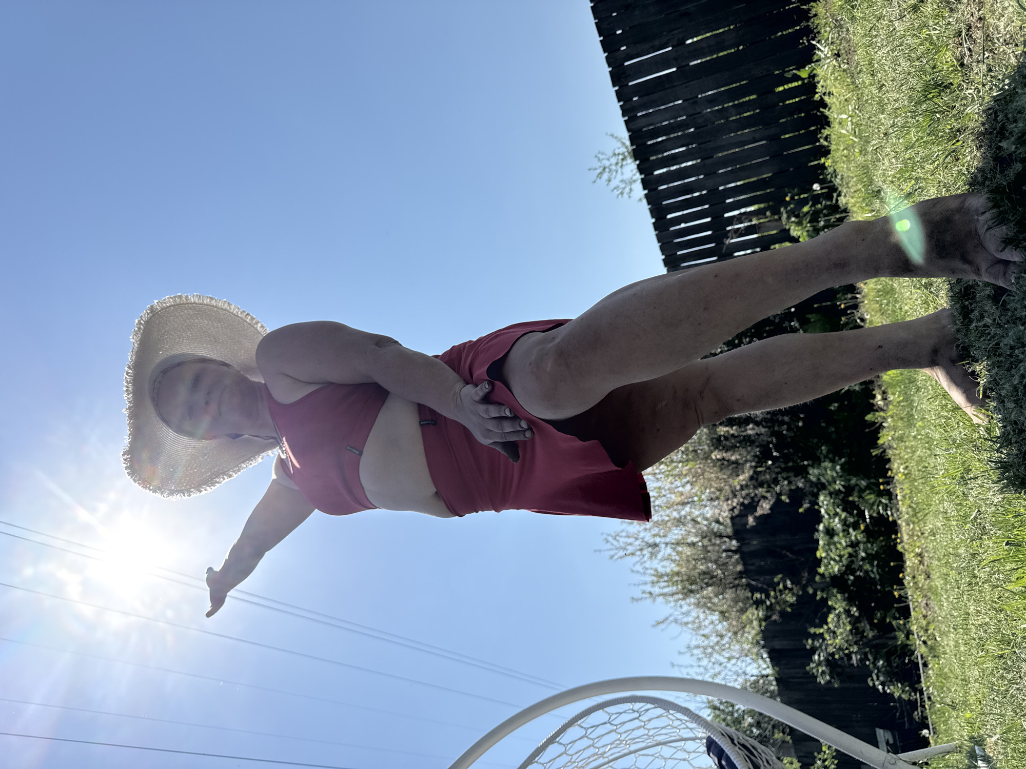 A medium-shot photograph depicts a woman wearing a wide-brimmed hat and a red swimsuit, standing outdoors. The environment includes a bright blue sky, green grass, a dark wooden fence, and possibly a partial structure of what appears to be a net. The photograph appears to have been taken from a low angle and is oriented sideways. The location is not specified.