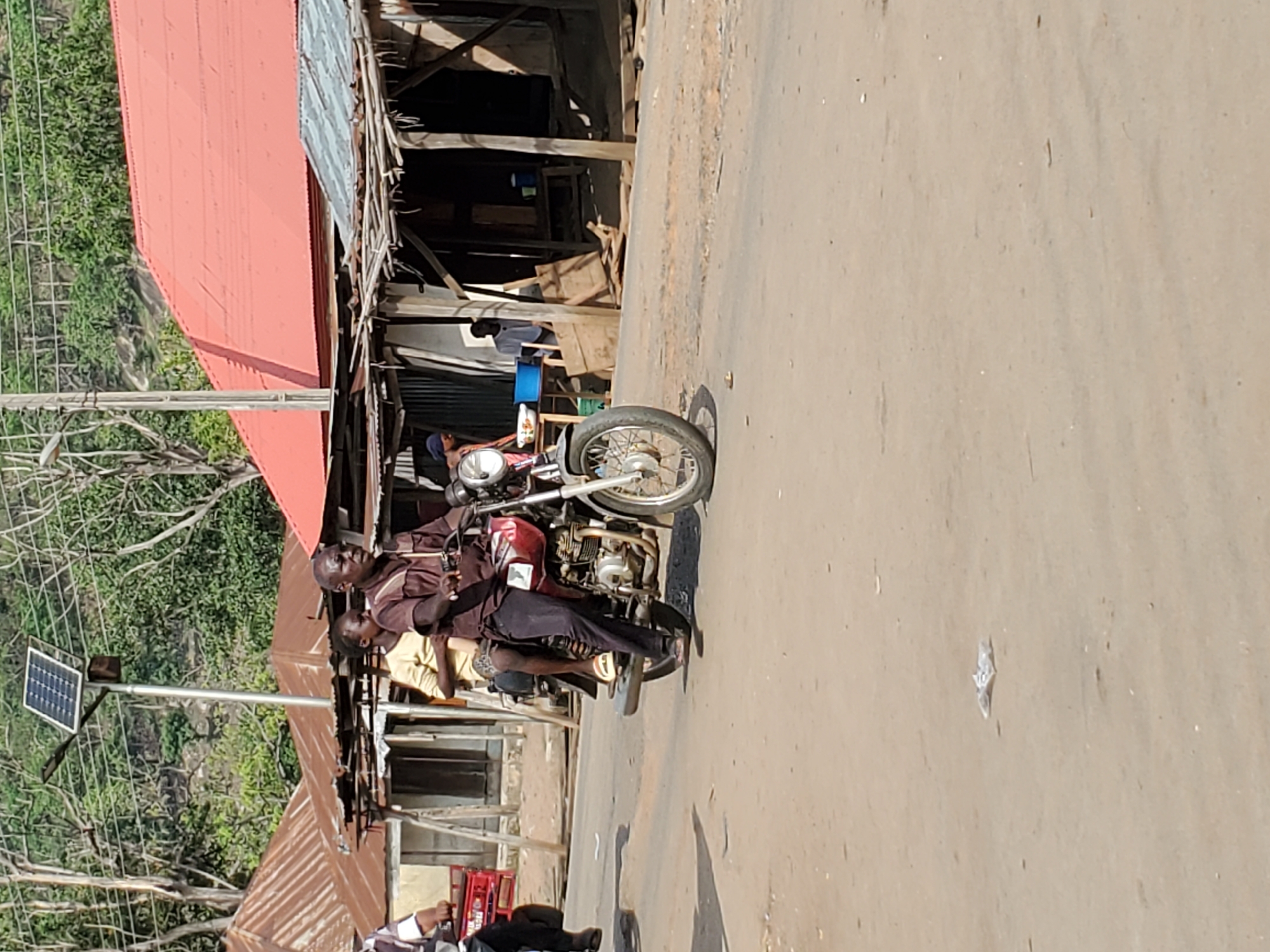 This image, likely taken in Mararaba, Nigeria, captures a daytime street scene with several key elements. The main subject is a person seated on a maroon motorcycle, positioned on an unpaved, light brown dirt road. The rider wears a dark brown long-sleeved shirt and dark pants, with sandals visible on their feet. Another individual, partially obscured and wearing lighter-colored clothing, is seated behind the driver on the motorcycle.
The setting appears to be a semi-urban or rural area. To the right of the motorcycle, several simple buildings with corrugated iron roofs are visible. Some roofs are painted red, while others are grey/silver. The structures feature exposed wooden support beams. In the background, lush green foliage and trees suggest a natural, vegetated environment, with what appears to be a rocky outcrop in the distance.
On the left side of the road, a modern solar panel is mounted atop a tall, slender metal pole, indicating a solar street light or power source. Nearby, a wire fence or utility lines run parallel to the road. The bright sunlight, casting subtle shadows, indicates it is daytime with clear weather. No discernable text is visible on any objects in the image. The overall scene depicts a typical daily moment in a local community.