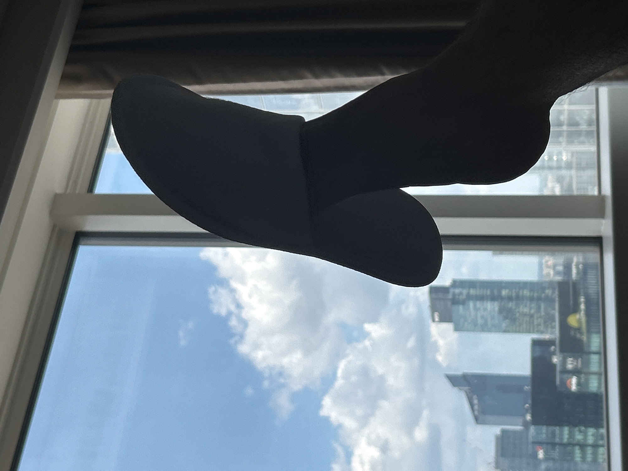 A silhouetted slipper-clad foot hovers in front of a window. The view through the window reveals a cityscape with modern high-rise buildings, indicative of a major urban center. The sky is partly cloudy. Based on the architecture and available information, the location is likely Warsaw, Poland.
