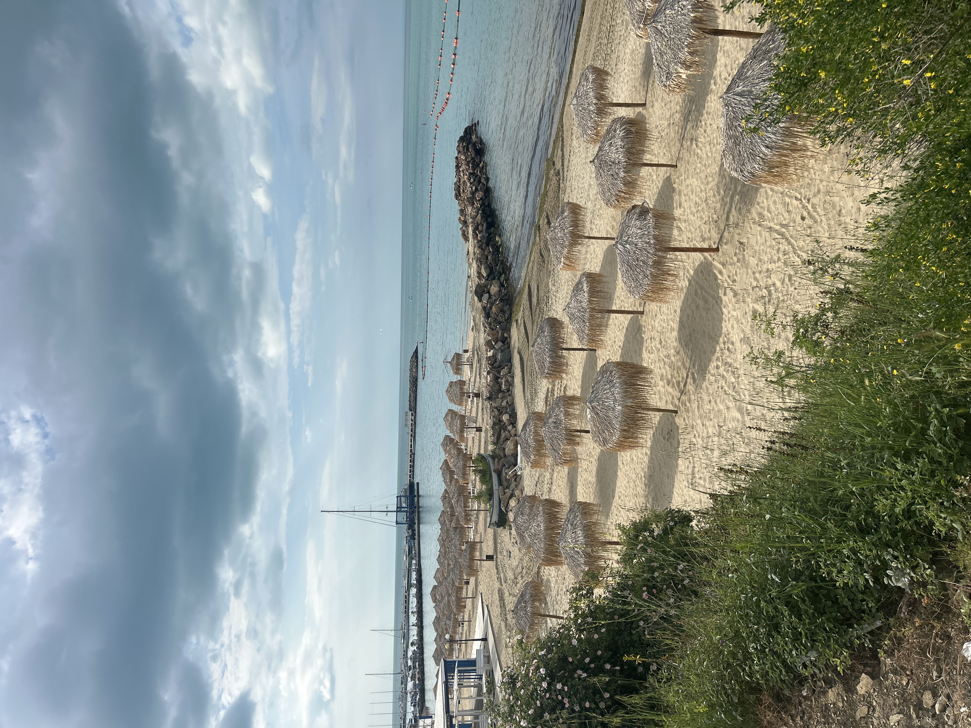 The image shows a beach scene in Sveti Vlas, Bulgaria. Rows of thatched umbrellas line the sandy beach. To the left is the sea, with a breakwater made of rocks and a pier visible in the distance. A string of orange buoys floats in the water. The sky is overcast with gray clouds. Green vegetation borders the beach on the right.