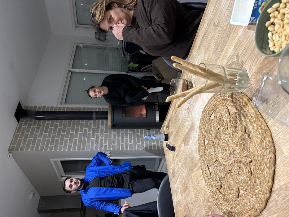 Three people are in a room. Two are sitting at a wooden table laden with breadsticks, peanuts, and a decorative place mat. One individual wears a brown sweater, while the other has on a bright blue jacket. A third person stands near a fireplace, wearing a black outfit. The room is lit by natural light coming through windows. The walls are white, and one has brick accents.