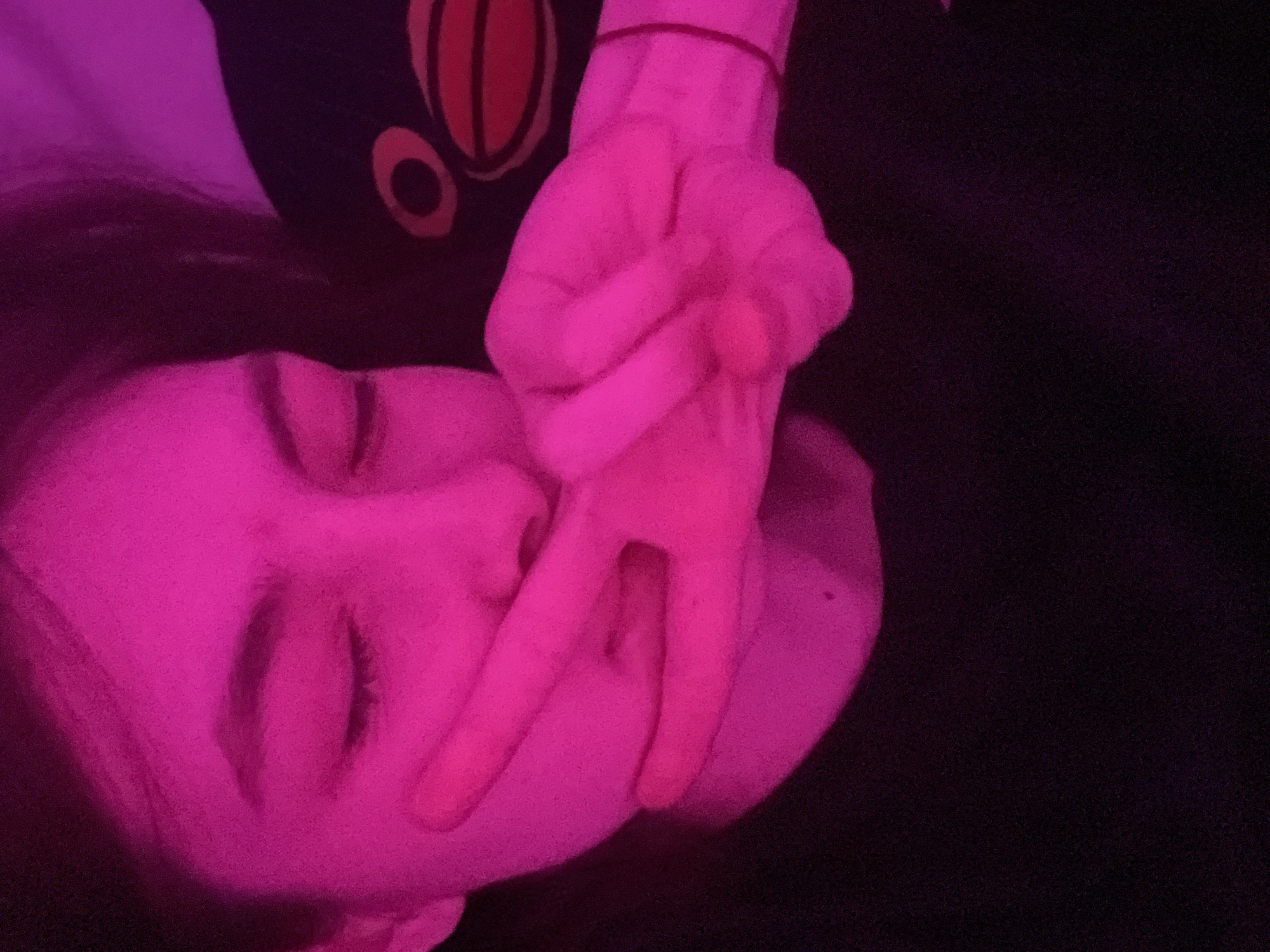 A close-up image, lit by a pink light, shows a person with their eyes closed, laying down. Their left hand is raised with two fingers extended, creating a V shape. To the top-left of the person's head, a dark object with a red and black pattern is visible. The background is predominantly dark. The location is not discernible from the image.