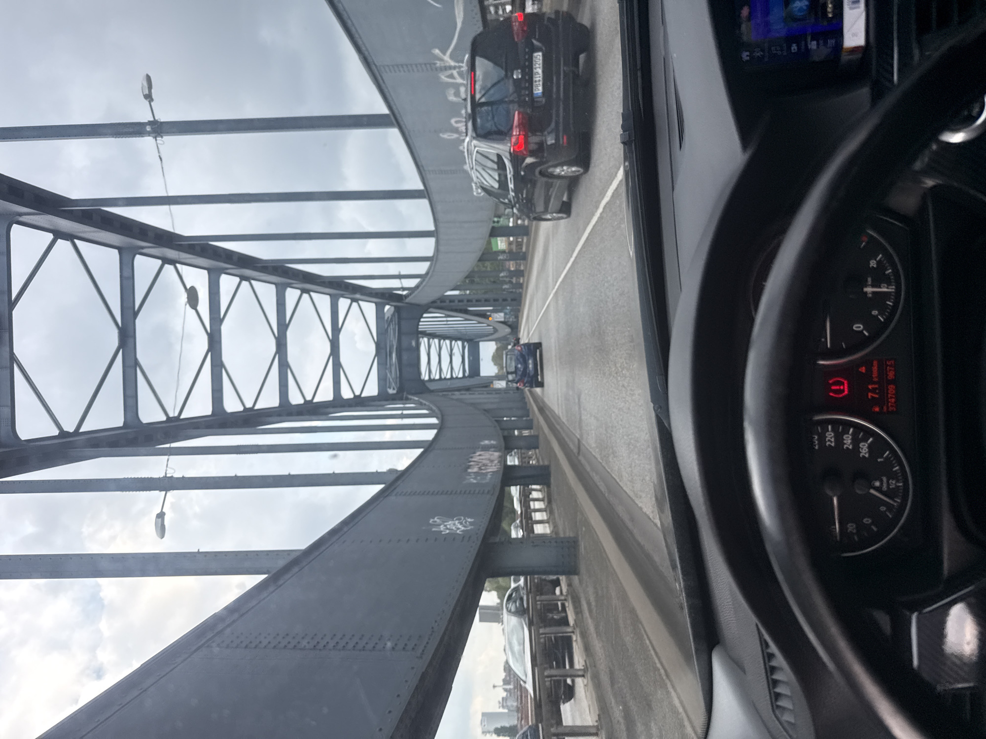 The image shows a view from the driver's seat of a car traveling across a large, gray steel truss bridge over the Elbe river in Hamburg, Germany. The sky is overcast. The car's dashboard and steering wheel are visible in the foreground, with a speedometer showing a speed of approximately 60 km/h. Several other vehicles, including a black SUV and a blue hatchback, are also on the bridge. The bridge structure, with its intricate metalwork and rivets, dominates the upper portion of the image. In the distance, city buildings are visible. The overall impression is one of urban transit on a cloudy day.