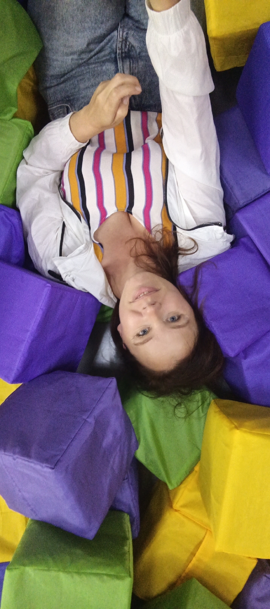 A young woman is lying on her back in a pile of large, colorful, soft cubes. She is wearing a white jacket over a striped shirt with vertical stripes of pink, yellow, black, and white. Her legs are bent and she is wearing blue jeans. Her left hand is resting on her abdomen. The cubes are predominantly purple, green, and yellow. The overall environment appears to be an indoor play area, possibly a ball pit filled with foam cubes. The image quality suggests it was taken with a mobile phone camera and is a selfie. The location is identified as Chaikovsky, Russia.