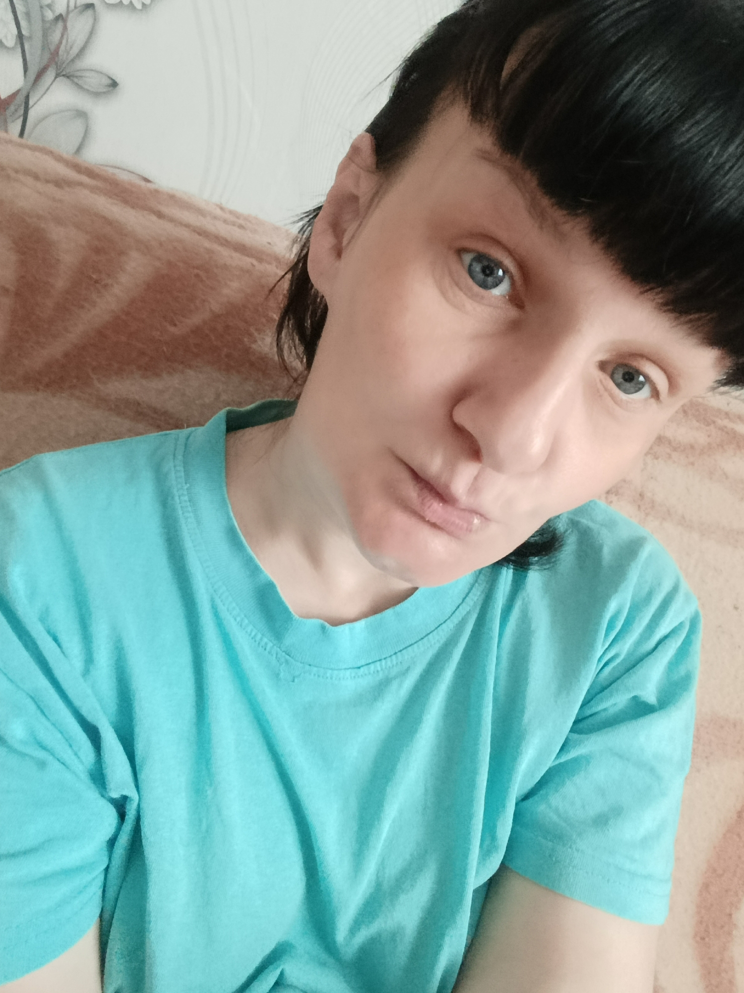 A close-up portrait of a light-skinned person with short, dark hair and blue eyes, wearing a bright blue t-shirt. The background appears to be an interior space, with a textured tan surface visible behind the person, potentially a couch or other upholstered furniture. A floral-patterned wall covering can also be seen in the background. There is no explicit evidence to confirm the location as Фоки, Russia.