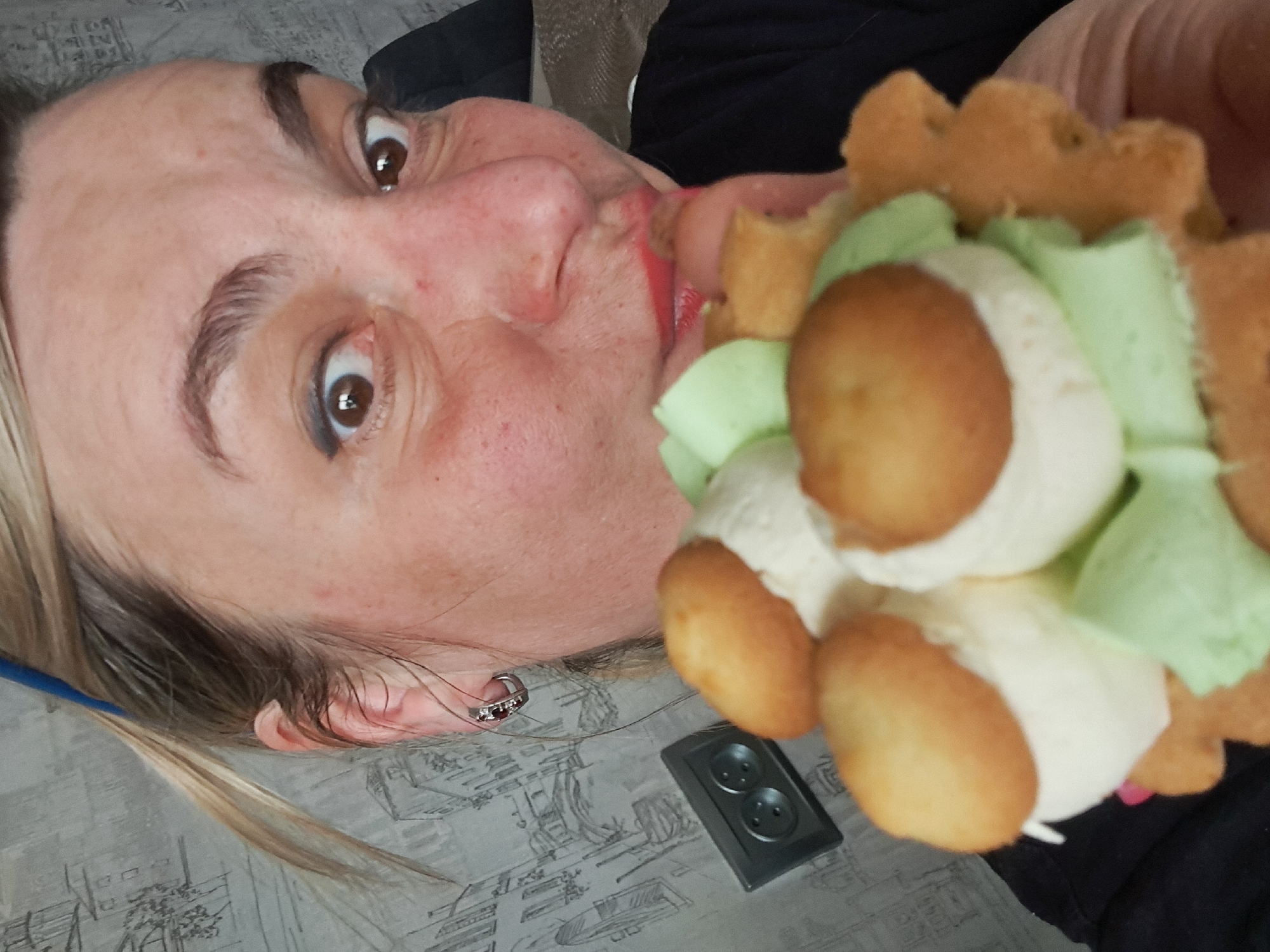 A woman is pictured close-up, her face tilted towards the camera, with an open mouth suggesting she is eating or about to eat a pastry. The pastry is decorated with brown puffs, white cream, and light green accents. She has light-colored hair, fair skin with freckles, and is wearing red lipstick and an earring. An electrical outlet is visible near her ear on a patterned surface. The image does not provide information about the location of Elektrostal, Russia.