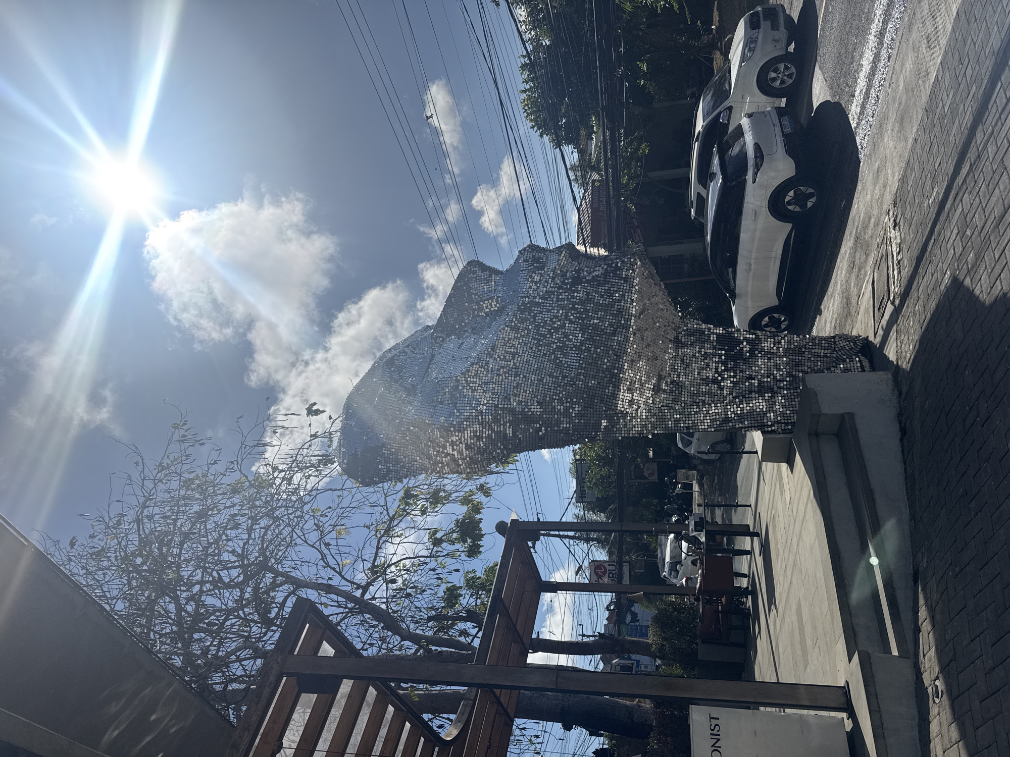 A large, abstract sculpture covered in shimmering silver sequins, resembling a human face or head in profile, dominates the foreground of this image. The sculpture is positioned outdoors, suggesting it's a public art installation or part of a commercial establishment. Behind the sculpture, a bright sun shines intensely in a blue sky with scattered white clouds, casting strong shadows and indicating it is daytime, likely mid-morning or afternoon given the angle of the sun's rays.
To the right of the sculpture, a paved sidewalk runs alongside a street where two white cars are parked. The street appears to be lined with trees and buildings, hinting at an urban or commercial setting. Overhead, a tangle of electrical wires crisscrosses the sky, a common sight in many urban environments.
In the lower left of the frame, parts of a wooden structure, possibly a pergola or seating area, are visible, suggesting an outdoor seating area associated with a cafe or restaurant.
The overall mood is bright and sunny, with the reflective nature of the sculpture adding a dynamic and eye-catching element to the scene. The location is Kuta Selatan, Indonesia. No people are clearly visible in the image, and no text is legible within the frame.