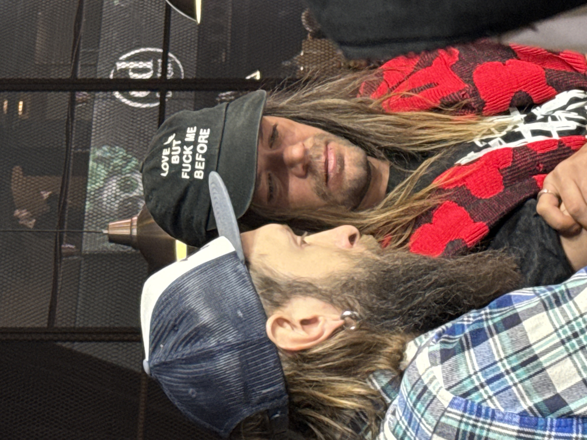 Two people are laying close together. The person on top is wearing a black baseball cap with the phrase "Love But Fuck Me Before" in white lettering. They have long, light brown hair and are wearing a black shirt covered by a red and black patterned garment. The person beneath them is wearing a blue and white baseball cap and a blue and white plaid shirt. They have a piercing in their ear. The background contains a black metal grate and other unidentified objects. The location and city of Khimki, Russia cannot be determined from the image.