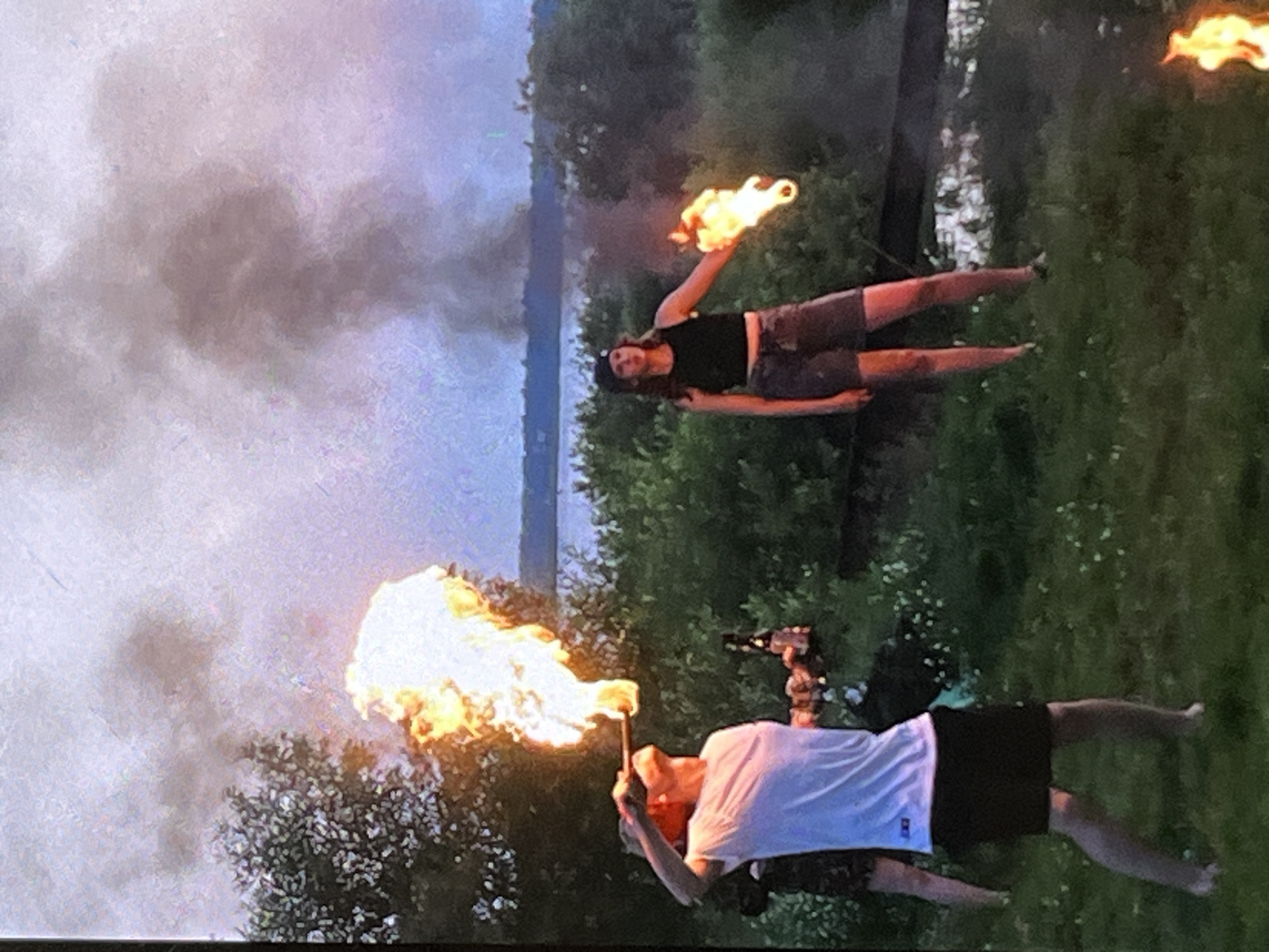 In a grassy outdoor area, possibly a park or backyard, near a body of water at dusk, two individuals are engaged in fire performance. On the left, a person in a white t-shirt and black shorts is blowing a large plume of fire from their mouth, holding a bottle in their left hand. On the right, a person wearing a black tank top and shorts holds a flaming object in their outstretched right hand. The background features dense green foliage and a hazy sky with smoke rising in the distance. The overall scene suggests a performance or recreational activity involving fire.