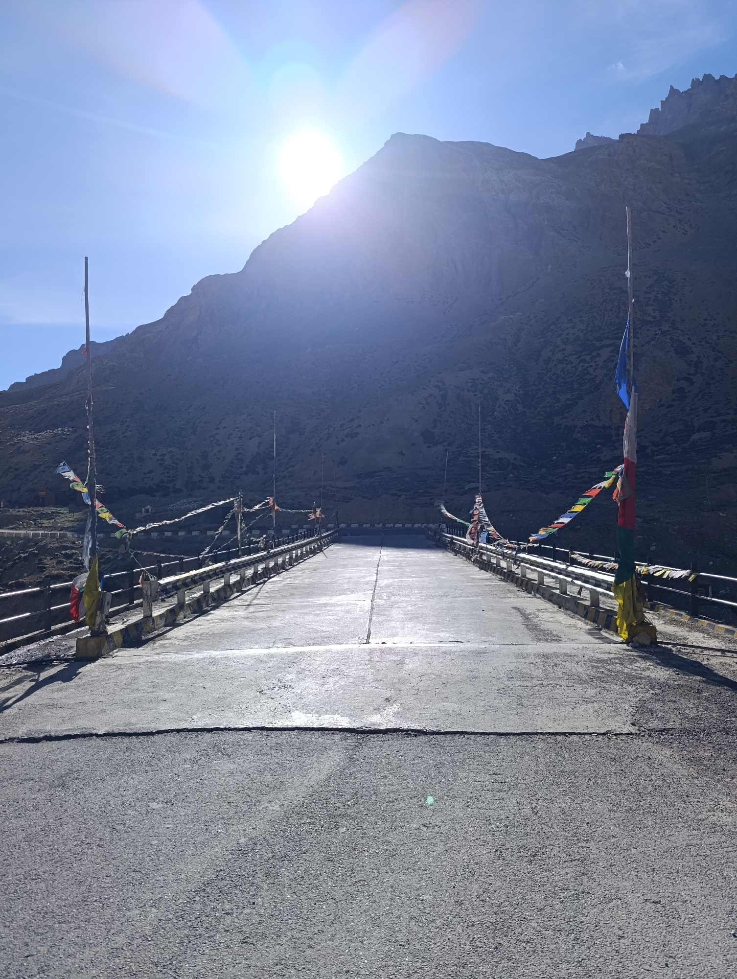 The image shows a bridge with prayer flags, leading towards a mountain under a bright sun and blue sky. The bridge is made of concrete and asphalt, with metal railings on either side. The prayer flags, colorful and fluttering, are tied to poles on the bridge and strung across it. In the background, a large, rugged mountain dominates the landscape, its slopes covered in rocky terrain. The sun is high in the sky, creating lens flare and casting long shadows. The overall impression is one of a remote, scenic location, likely in a mountainous region. The location is described as Dhar Chharp Samba, India. There are no people visible in the image.