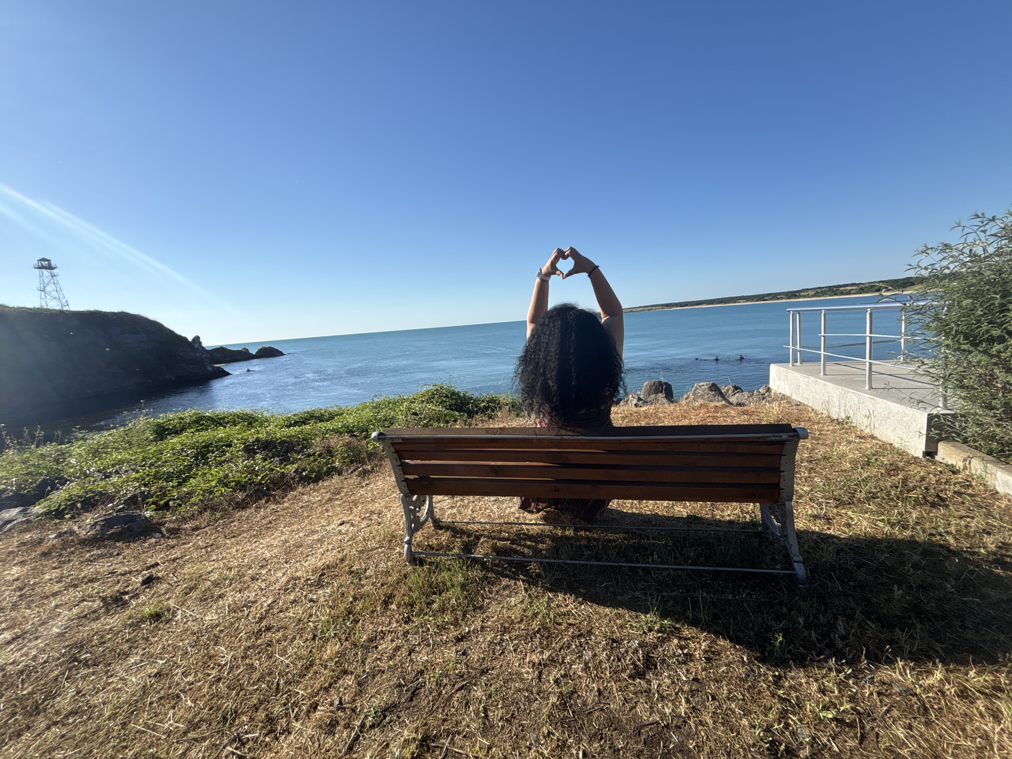 A person with long, dark, curly hair is seated on a wooden bench facing away from the camera. Their arms are raised, and their hands are forming a heart shape against the clear blue sky. The bench is situated on a grassy, dry hillside overlooking a calm blue sea. In the distance, a rocky shoreline with a small tower-like structure on a cliff is visible to the left. The sea extends to the horizon, meeting the sky under a bright sun. To the right of the bench, a concrete pathway with a white railing runs alongside a patch of green foliage. The location appears to be the coast of Rezovo, Bulgaria.