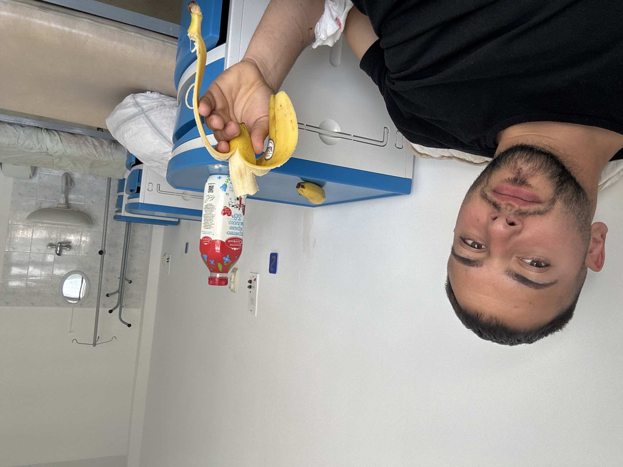 The image shows a man holding a partially peeled banana in his right hand. He is wearing a black t-shirt. The background appears to be a hospital room with medical equipment, including a bed and an IV stand. There is a juice box with a red top and pictures of blueberries and strawberries. There is a small, unpeeled banana on a shelf. The location may be a hospital in Sliven, Bulgaria based on the markings on the juice box.