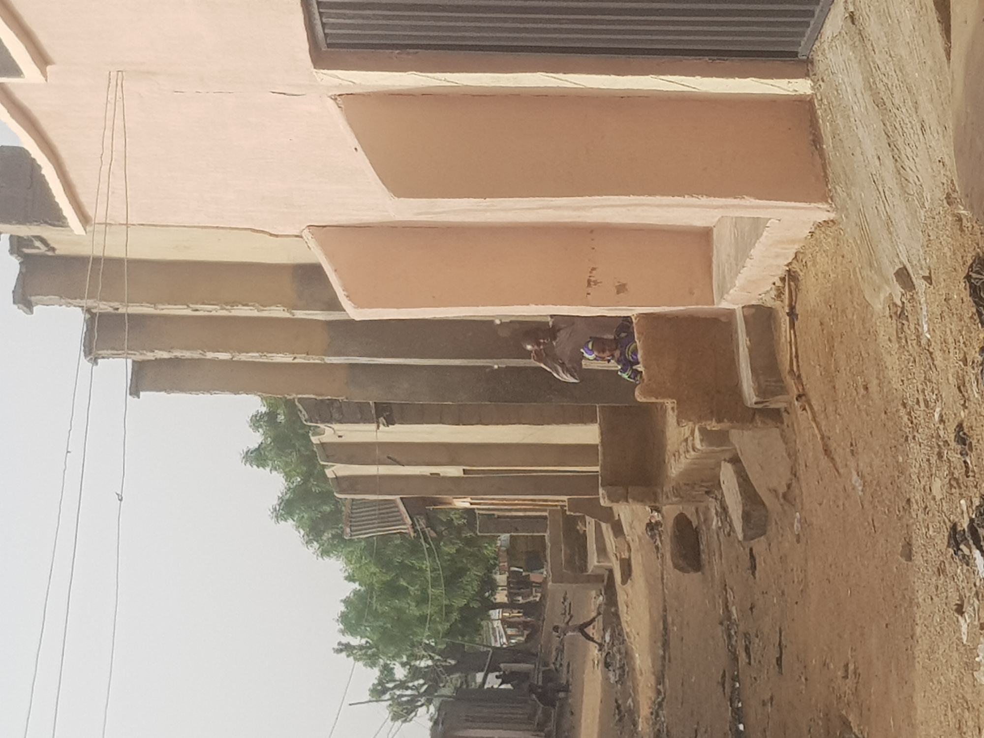 A daytime shot in Dutsinma B, Nigeria, showcases a narrow street lined with buildings. The structures are constructed from light brown materials, possibly mud or adobe brick. The camera angle, positioned from a low point and tilted, captures the underside of the buildings' overhangs, which create a rhythmic pattern of projecting ledges.
A figure, presumably a resident, is partially visible under the overhang. The person is wearing what appears to be a patterned shirt.
Overhead power lines are visible, and a few trees with green foliage are seen. The ground is uneven and covered with debris, indicative of an unpaved street. The lighting is diffused, suggesting an overcast day.