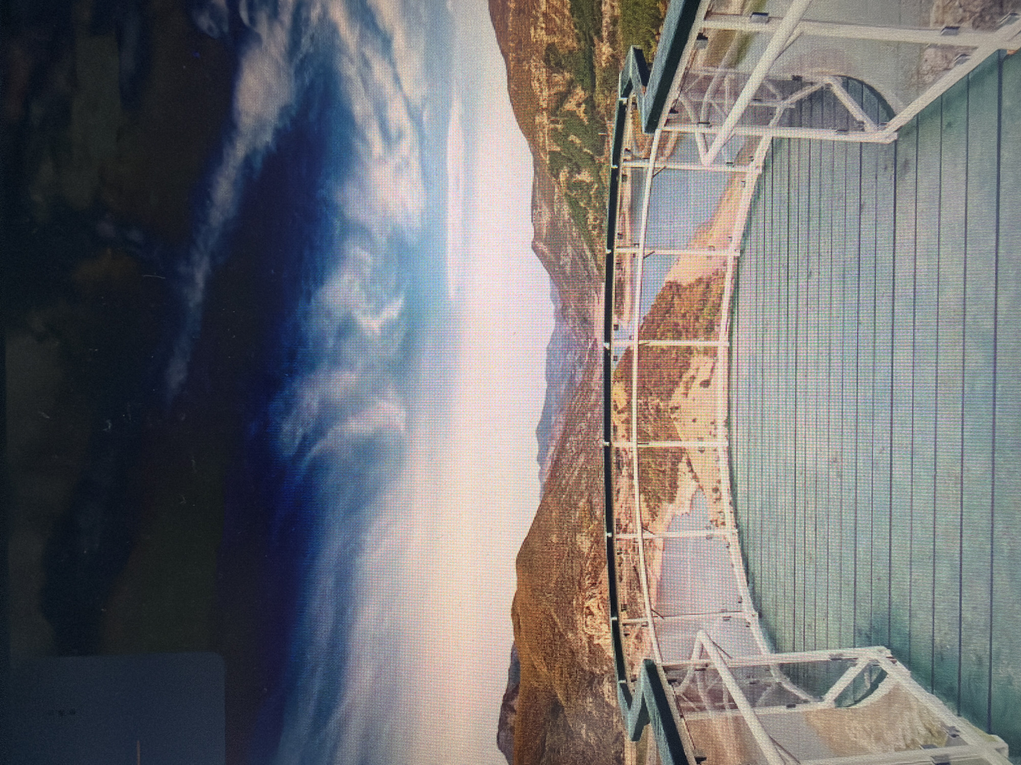 The image shows a view from a circular observation deck looking out over a body of water winding through a valley of rolling, arid hills under a dramatic sky. The deck has a light green wooden floor and white railings with glass panels. In the distance, the hills are covered in dry vegetation and exposed rock formations. The sky is a mix of deep blues, blacks, and whites, with streaks of clouds suggesting movement. The scene is a landscape vista, likely a dam or a large reservoir.