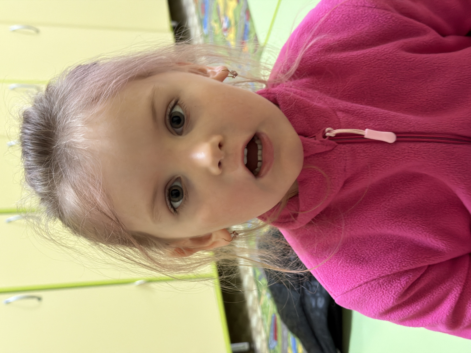 A close-up, sideways portrait of a young child, likely a girl, is captured indoors. The child has fair skin, light brown or blonde hair, and light-colored, wide-open eyes. Her mouth is open, revealing her teeth, suggesting an expression of surprise, curiosity, or engagement. She wears a bright pink fleece jacket with a visible zipper and a white and pink zipper pull. Small, gold-colored hoop earrings are visible in her ears. The background features light green cabinets or furniture with silver handles, indicating an indoor setting, possibly a playroom or classroom. A colorful, patterned surface, likely a rug or mat, is partially visible in the upper right portion of the background. The lighting is bright and even. No text is visible in the image. This scene takes place in Chaikovsky, Russia.