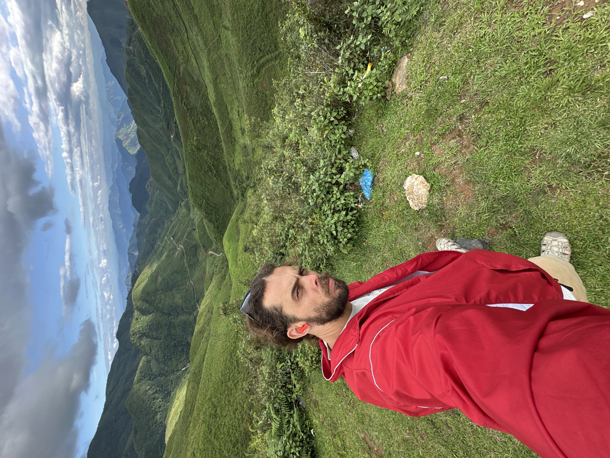 The image captures a selfie of a male individual on a grassy slope overlooking a vast mountainous landscape in Xín Vàng, Vietnam. The man, appearing Caucasian with brown curly hair, a beard, and sunglasses resting on his head, wears a bright red jacket with white trim, a grey t-shirt underneath, light-colored pants, and light-colored open-toed hiking sandals. Orange earplugs are visible in both ears. He looks directly at the camera with a neutral to stern expression.
The environment is characterized by numerous layers of steep, undulating green mountains and valleys, heavily covered in dense vegetation. A winding road is discernible traversing through the valleys and along the lower slopes of the mountains in the midground. The sky above is partially covered with large, grey clouds, interspersed with clear blue patches and smaller white clouds. In the foreground, immediately surrounding the man, the ground is covered in grass, scattered rocks, and patches of darker green bushes and ferns. Visible among these bushes, to the right of the man, is miscellaneous litter, including a blue plastic bag, a white plastic item, and a yellow wrapper.