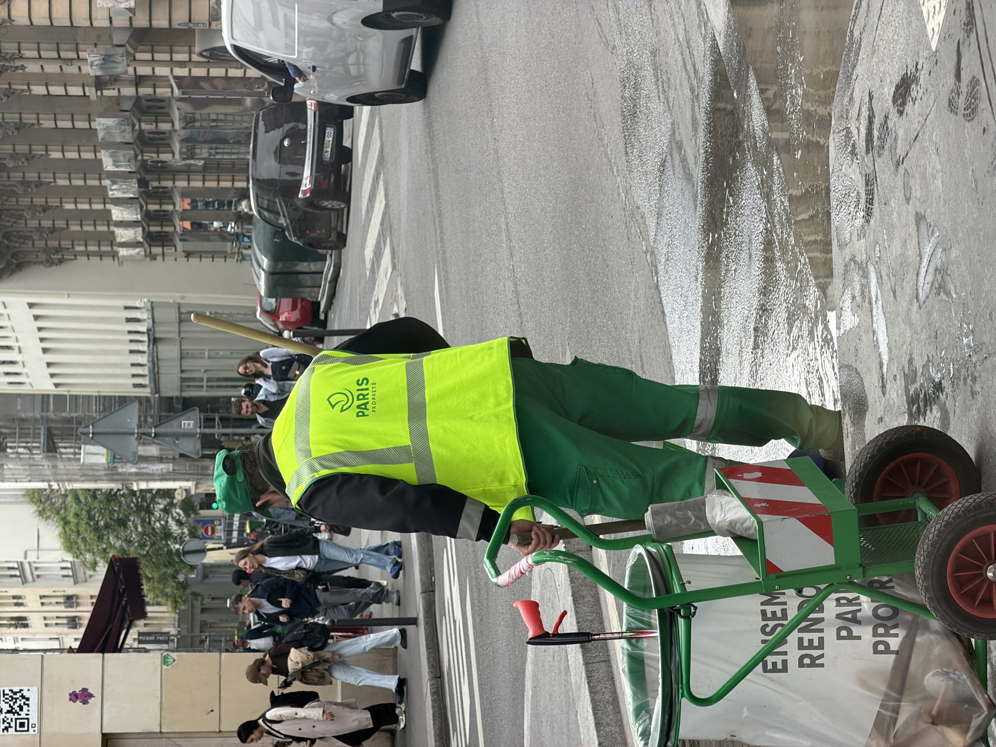 A sanitation worker in Paris, France, wearing a bright yellow vest with the "Paris Proprete" logo and green pants, stands on a sidewalk and pulls a green cart with red wheels. The cart contains trash and the phrase "ENSEMBLE RENDONS PARIS PRO." printed on it. The worker is near a crosswalk on a wet street, with cars passing by. Pedestrians walk behind the worker on the sidewalk. Buildings are visible in the background.