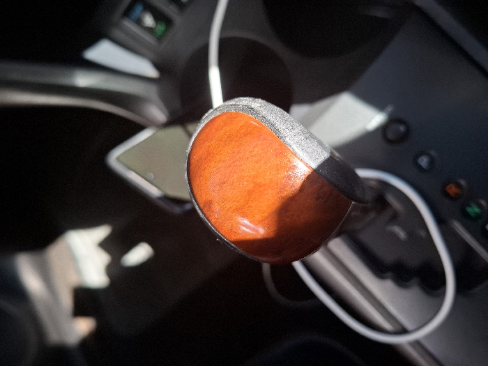 The image shows the gear shifter of a car, likely located in a vehicle in Gitaru, Kenya, at the coordinates provided. The gear shifter is in the park position, with the shifter knob visible. The knob is brown and appears to be made of wood. It is surrounded by the black interior of the vehicle. The shifter base is silver with a black outline. A white cord is wrapped around the shifter base, suggesting it is connected to a device. The image also shows a part of the car's console, with several buttons and a control panel in the background. The lighting is bright, suggesting it is daytime and the car is parked. No people are present in the image.