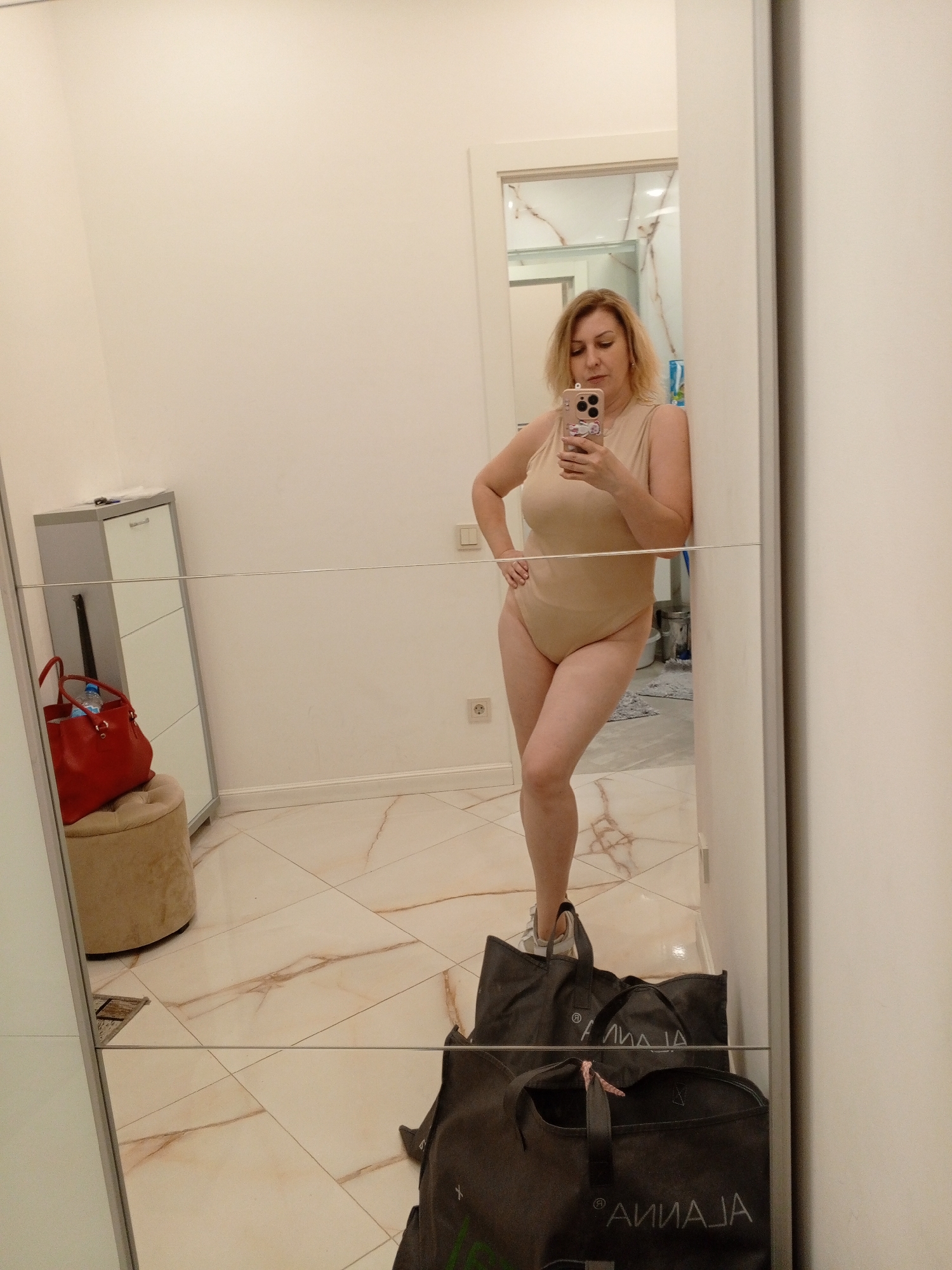 A woman stands in what appears to be a hallway or dressing area, taking a selfie in a full-length mirror. She is wearing a beige sleeveless bodysuit and white sneakers. Her blonde hair is styled in a bob, and she has a neutral expression.
Behind her, reflected in the mirror, is a shoe cabinet with a red handbag and a water bottle on top. To the side, a round, beige ottoman is visible. The floor is tiled with a marble-like pattern. Several large, dark shopping bags are on the floor in front of the mirror.
The scene is indoors, with bright, artificial lighting. The overall impression is a casual, personal moment. No text is clearly visible in the image.