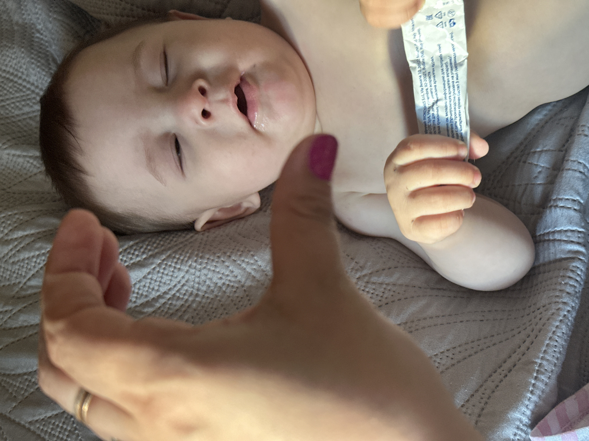 A medium shot shows a baby lying on a textured gray blanket. The baby's eyes are closed, and their mouth is slightly open, with some liquid visible. A hand with pink nail polish is in the foreground, positioned to the left of the baby's face, with the thumb and forefinger forming a heart shape. The hand is wearing a thin gold ring on the ring finger. The baby's chest and abdomen are visible, and they are holding a white tube with text printed on it in their hands. The background consists of the gray blanket. The scene is indoors, and the lighting appears natural.