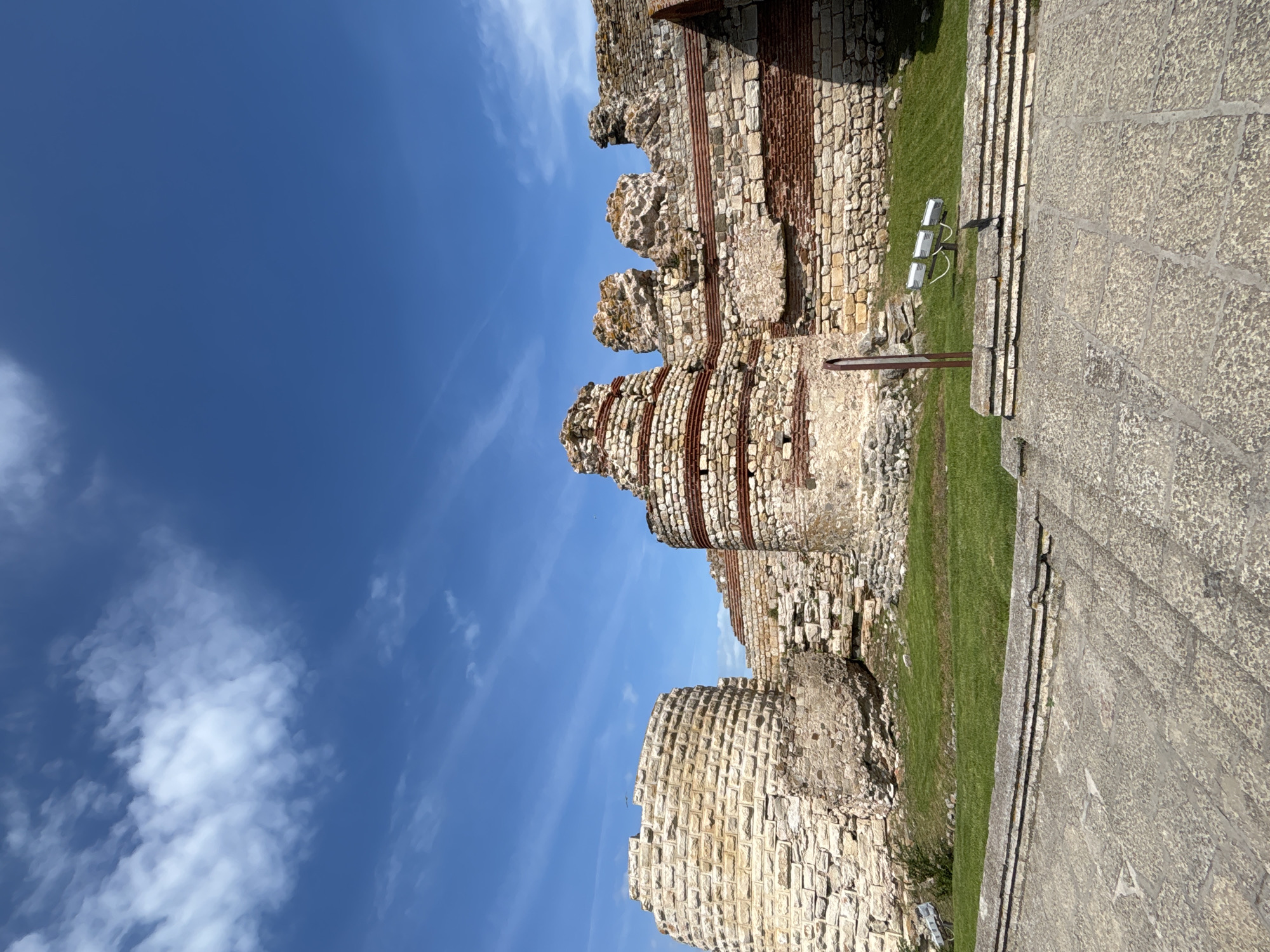 The image shows the ruins of ancient stone fortifications under a bright blue sky with scattered white clouds. The fortifications are made of weathered, light-colored stones with reddish-brown bands interspersed throughout. The structures are partially collapsed, revealing their layered construction and age. In the foreground, there is a paved area with large, irregular stone slabs, leading to a low stone wall. Behind the wall is a well-maintained patch of green grass. Three white spotlights on poles are visible on the grass, positioned near the ruins. To the far left, a portion of a cylindrical stone structure, appearing to be a tower or section of a wall, is prominent. The scene captures a historic landmark in Nesebar, Bulgaria, likely a part of the ancient city walls. There are no people visible in the image. The lighting suggests it is daytime, likely late morning or early afternoon due to the bright sun and shadows.