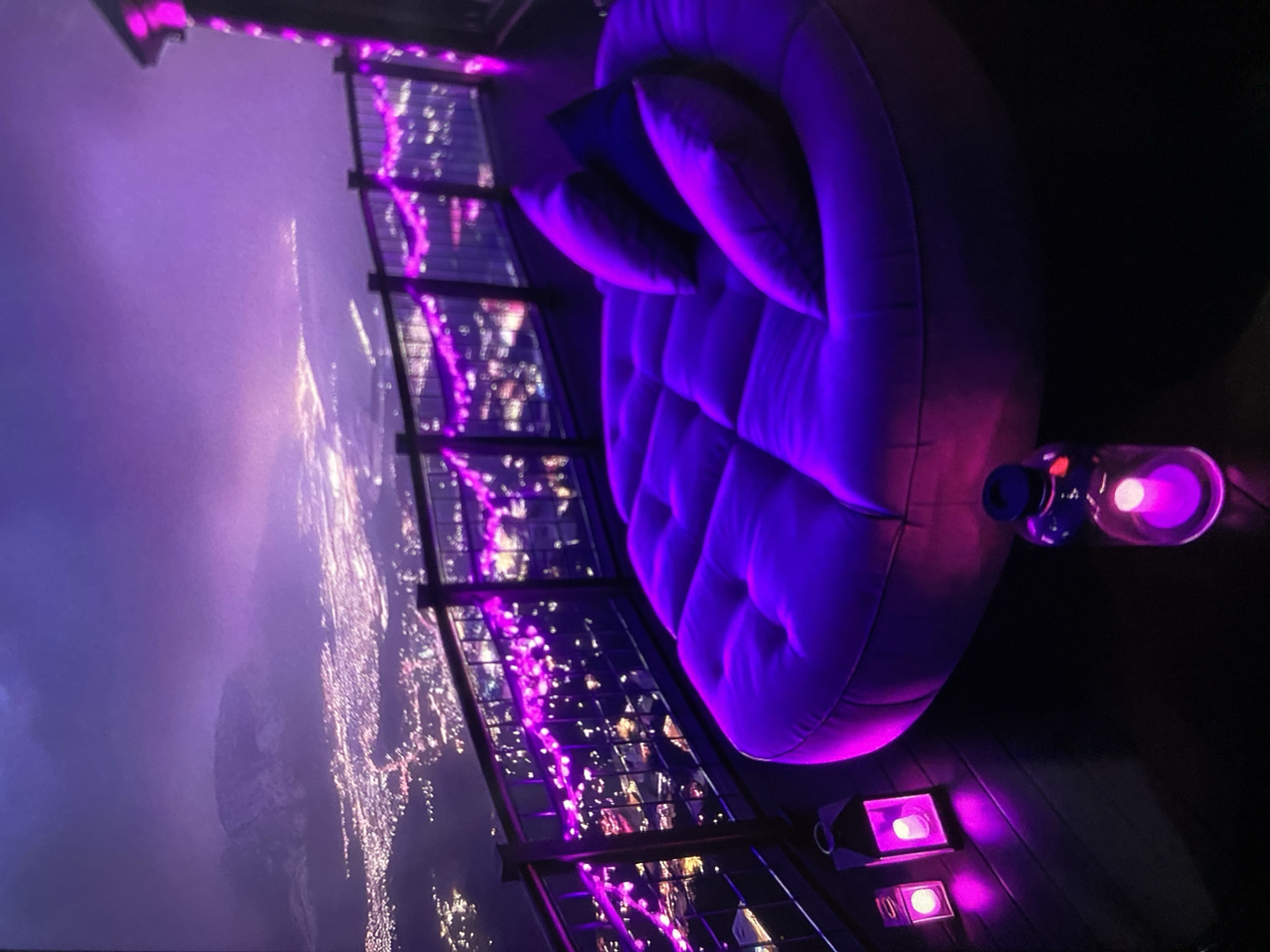 The image presents a nighttime scene from a high-altitude location, likely a rooftop or high balcony overlooking a city. The foreground features an oversized, round, upholstered sofa in a vibrant purple hue, adorned with darker-colored pillows. To the right of the sofa, there are several light sources emitting a similar purple light, including two spotlights and a decorative bottle-like lamp with a glowing element. The setting is further enhanced by string lights draped along the edges of the structure, also radiating a purple glow.
Beyond the structure, the city stretches out into the distance, its lights creating a sprawling urban landscape. A large landmass, possibly a mountain or significant terrain feature, can be seen on the left. It cannot be definitively stated that the image is of Lhokseumawe, Indonesia based on visual information alone.