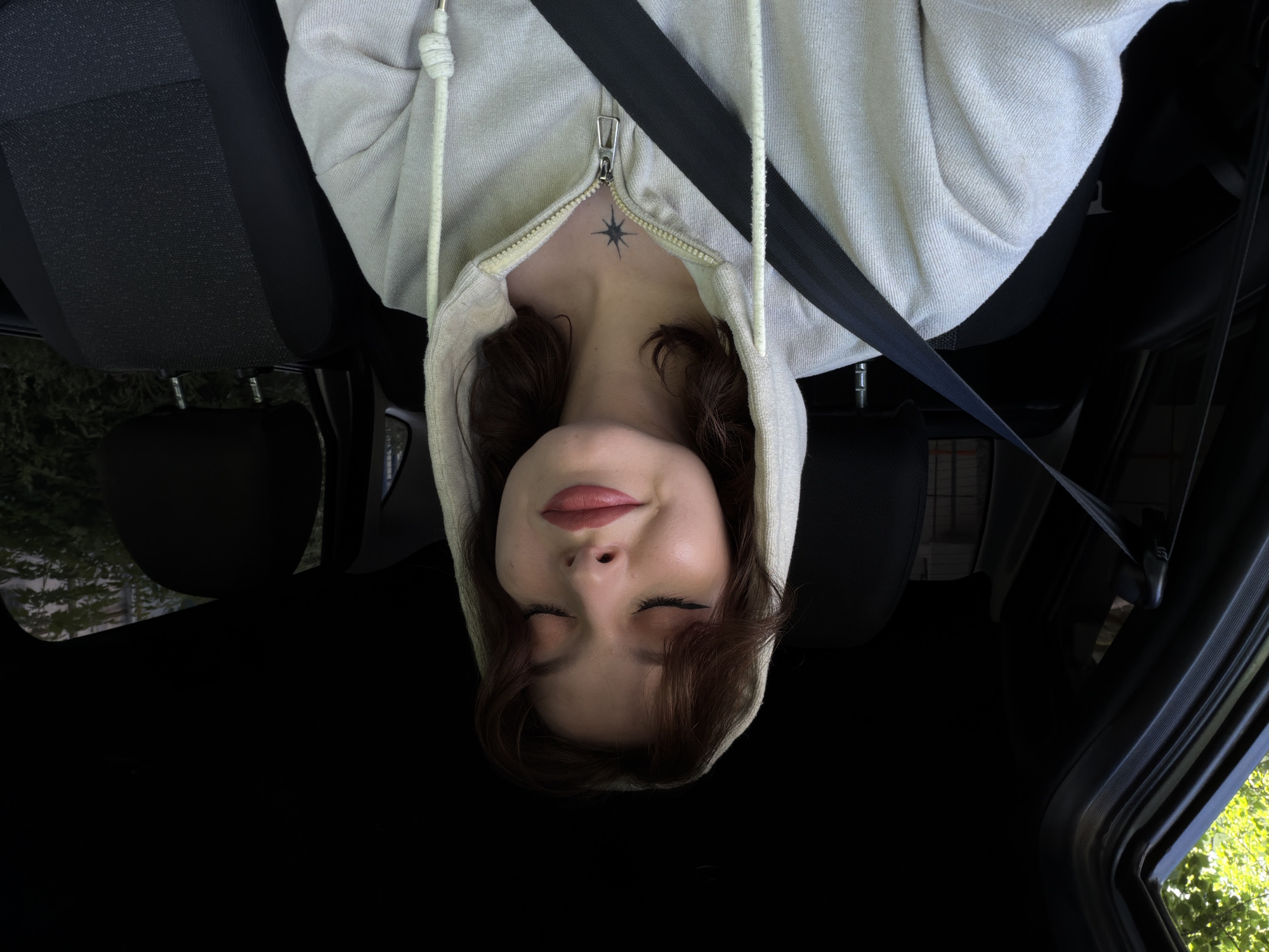 A young woman with brown hair and closed eyes is sitting in the driver's seat of a car, wearing a light-colored hooded jacket and a seatbelt. She has on makeup, including eyeliner and lipstick. A star-shaped tattoo is visible on her chest. The car's interior, including the seats and dashboard, is dark. The exterior, visible through the windows, shows green trees and foliage.
