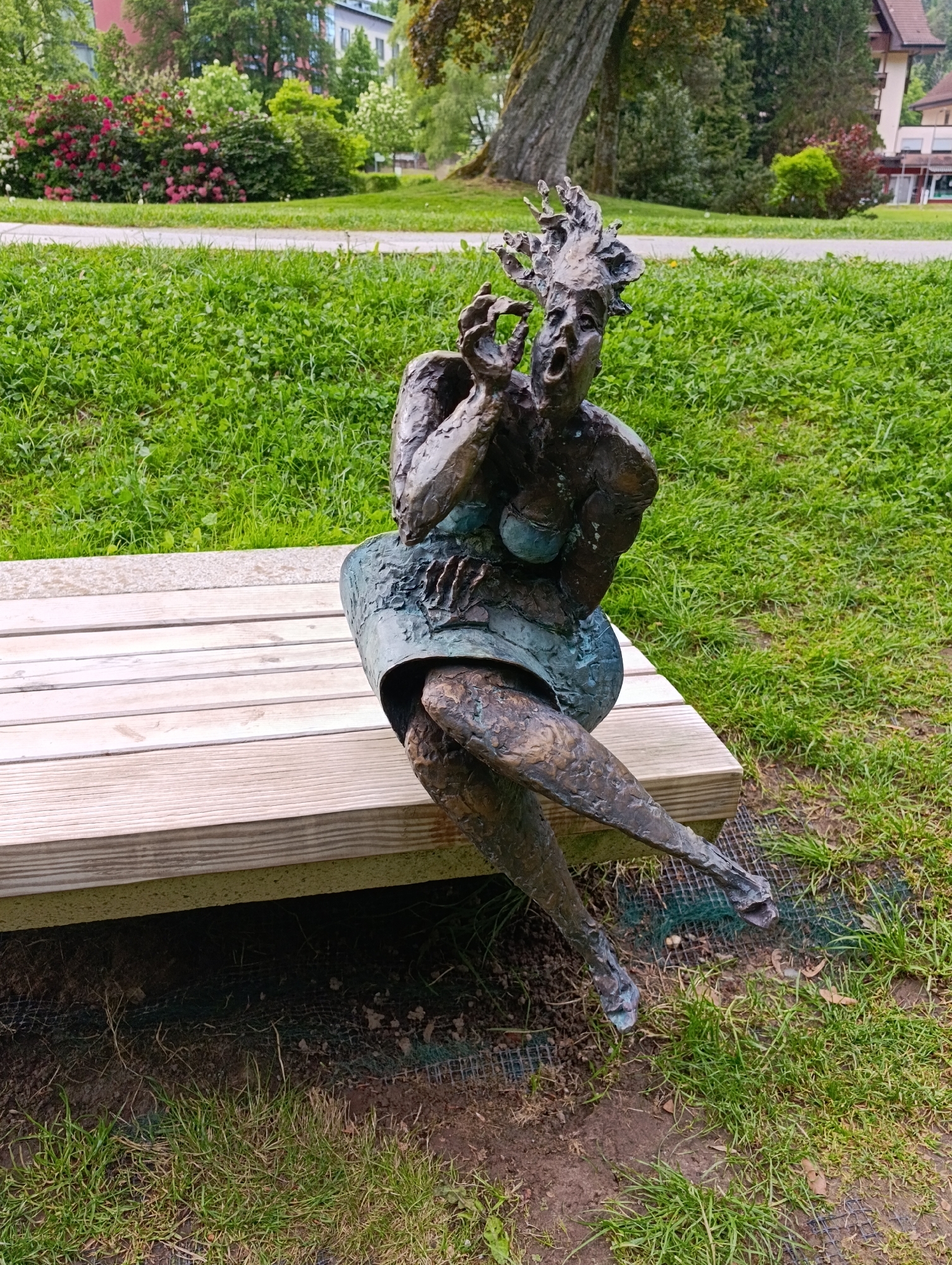 Here is a detailed description of the image:
The image features a bronze-colored sculpture of a woman sitting on a light wooden bench. The woman has an exaggerated facial expression, appearing to be screaming or expressing shock, with her mouth open wide and hands raised to her face. Her hair seems wild or unkempt, styled in a disordered manner. She wears what appears to be a short skirt or dress, and her legs are crossed with her feet slightly off the ground.
The bench is simple, made of light-colored wooden slats and placed on a grassy lawn. Behind the bench, there is a park-like setting with green grass, trees, and flowering bushes. The grass is neatly manicured, suggesting a well-maintained park. In the background, there are buildings, indicating this is likely an urban or suburban area.
The scene is located in the city of Unknown, Unknown. The environment seems peaceful despite the dramatic sculpture, suggesting that this piece of art is integrated into the urban landscape for public enjoyment.