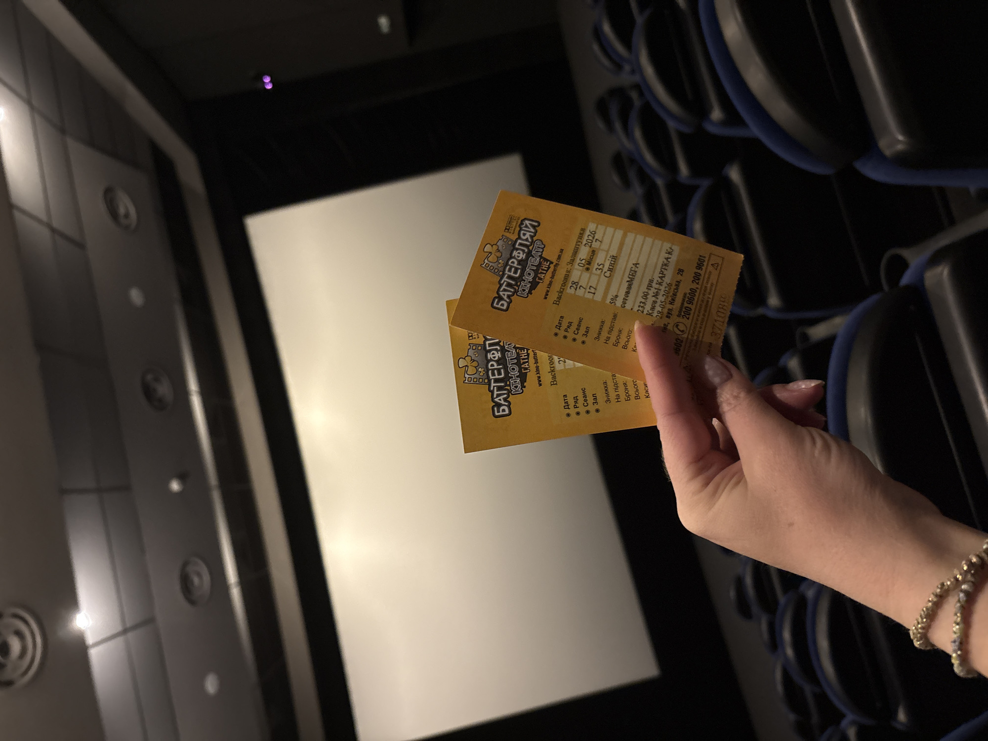 The image shows a hand holding two movie tickets for the "Баттерфляй Кінотеатр Гатне" (Butterfly Cinema Hatne), located at вулиця Київська, 28 (Kyivska street, 28) in Hatne, Ukraine. The movie being shown is "Backrooms: Залаштунки" (Backrooms). The tickets are for May 5, 2026, Row 2, Seat 7 in Hall 17 and the movie's showtime is 28:35. A discount is listed as 5% and the ticket price is 233.00 грн. In the background, a white movie screen is visible, along with rows of blue and black movie theater seats. The scene is dimly lit.