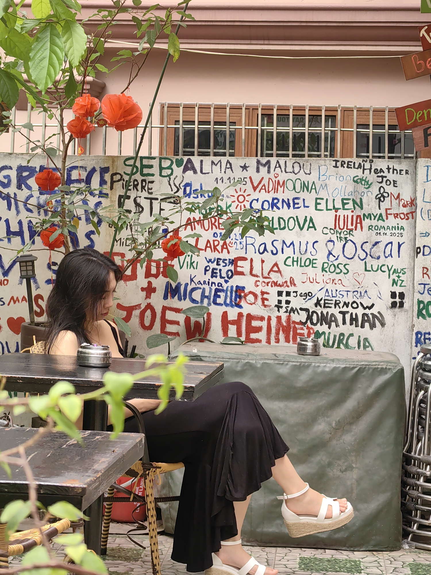 A medium shot shows a young Asian woman seated at a dark-stained wooden table in an outdoor café setting in Bai Chay Ward, Vietnam. She wears a black maxi skirt, a black top, and white platform sandals. A potted plant with vibrant red flowers and orange lanterns stands near her and adds color to the scene. The café's back wall is covered in graffiti. A covered table with a silver ashtray is visible in the background, along with stacked metal chairs. The image has a slightly blurred foreground due to nearby foliage.
