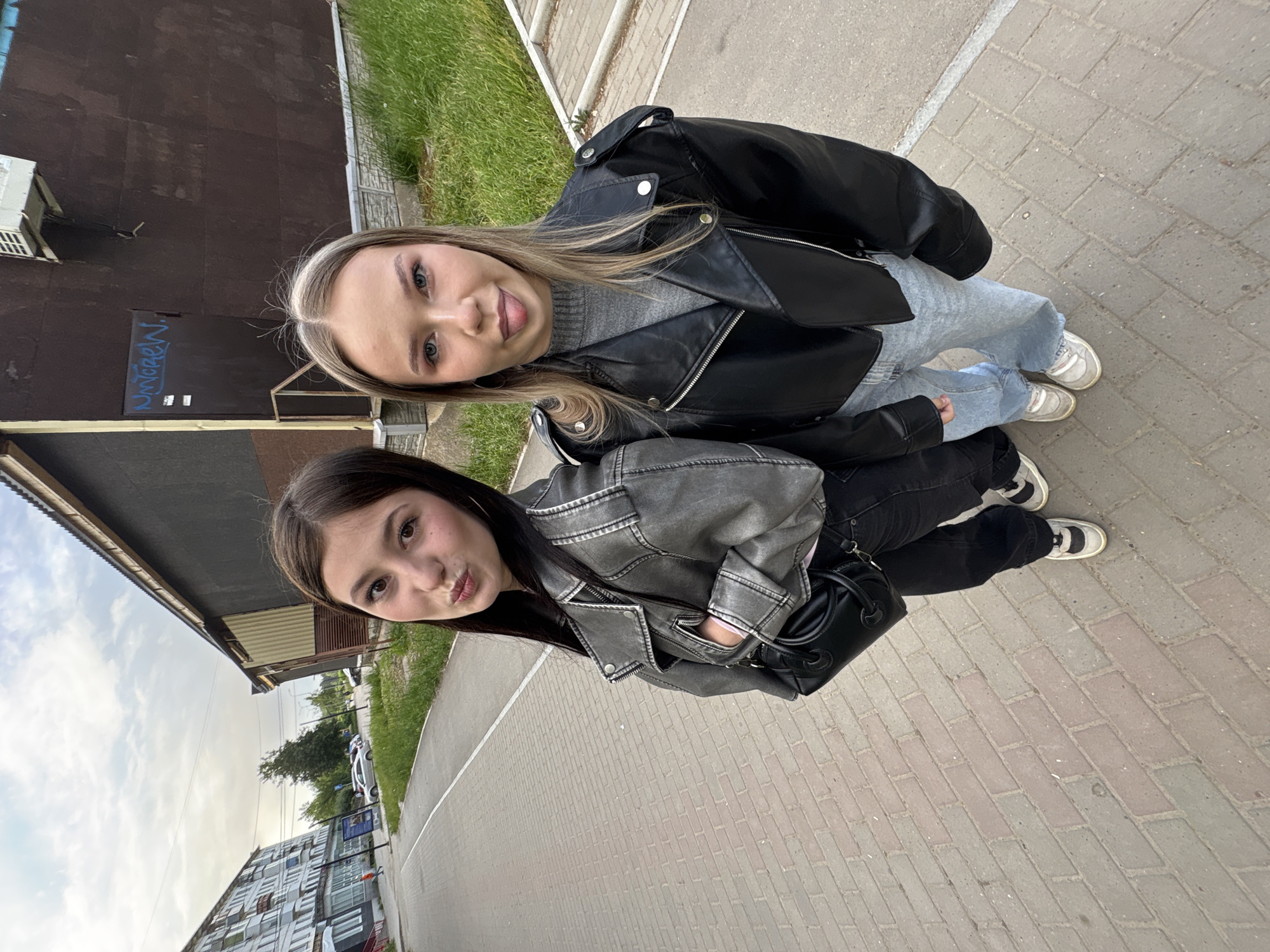 Two young women stand close together on a brick-paved walkway. The woman on the top, closer to the camera, has blonde hair and is sticking her tongue out. She wears a black leather jacket over a gray sweater and light blue jeans. The woman on the bottom has dark brown hair and is pouting slightly. She wears a gray leather jacket over a pink shirt and black pants. She is holding a small black purse. Both women are wearing sneakers.
The background includes a brown building with some graffiti, a grassy area, and a sidewalk. Across the street, there are apartment buildings and cars parked along the road. The scene appears to be in a Russian city, likely Chaikovsky.
