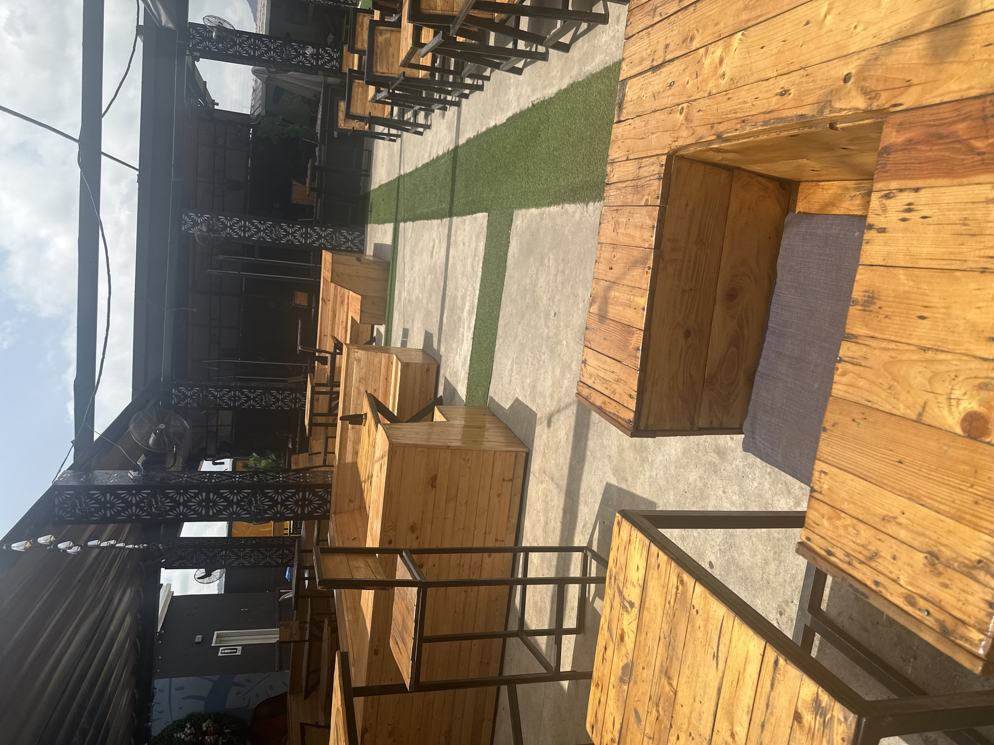 The image shows an outdoor seating area with wooden tables and benches. The furniture is made of light-colored wood and has a rustic design. Some tables are square, while others are rectangular. There are also some high stools with metal frames. The seating area is paved with concrete and has some patches of artificial grass. In the background, there is a covered structure with a black fabric roof, supported by metal poles. Decorative metal screens with floral patterns are visible on the sides of the structure. Two ceiling fans are mounted on the structure. The sky is partly cloudy. There are no people visible in the image. The scene appears to be a restaurant or a lounge area in Howayi, Nigeria, with the typical open-air setup often found in such establishments.