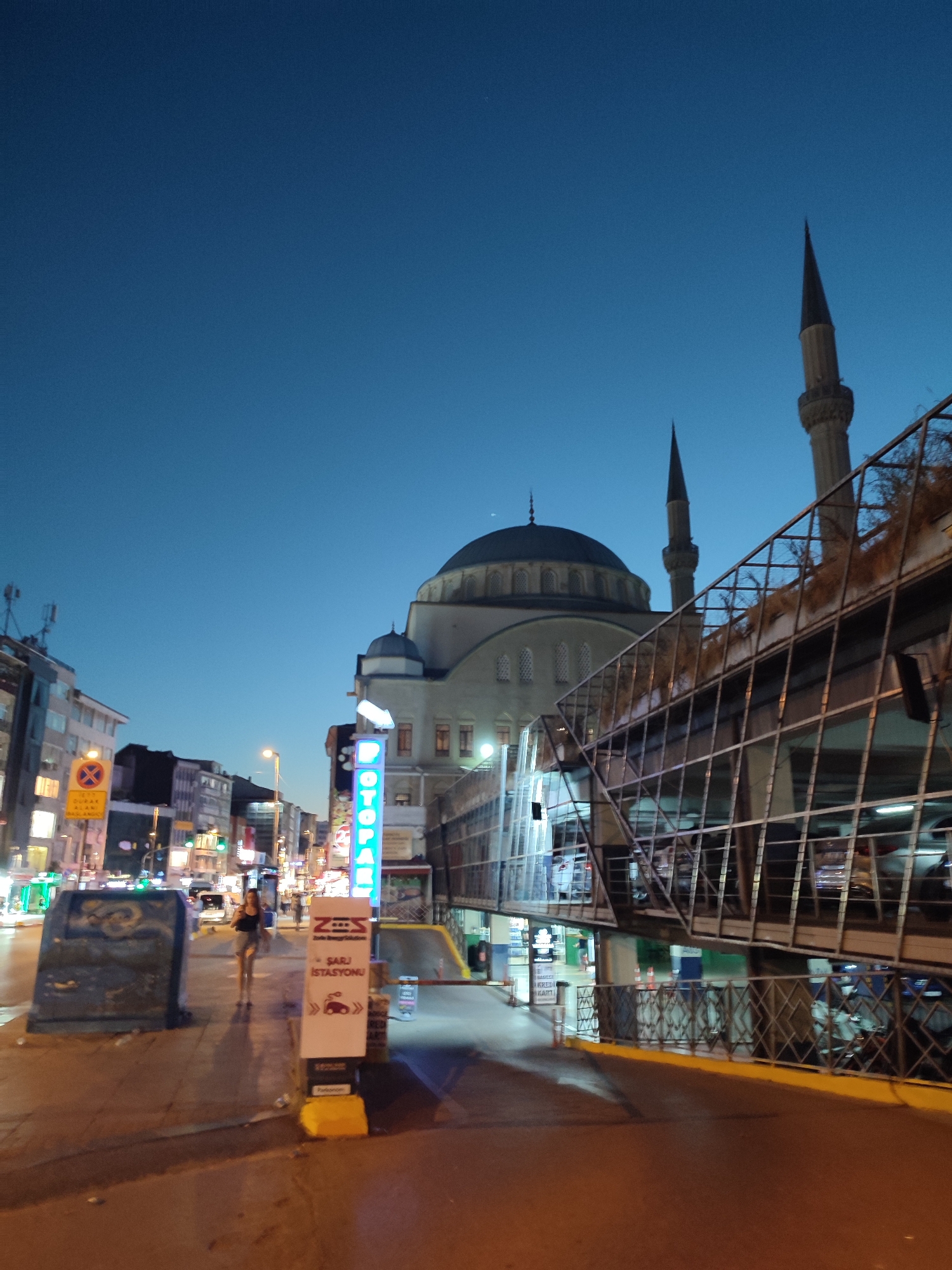 The image depicts a street scene in Kadıköy, Turkey, at dusk. Dominating the background is a large mosque with a prominent dome and two minarets, characteristic of Islamic architecture. In the foreground, a modern multi-story parking structure with a glass facade is visible, angled towards the viewer. The parking structure appears to be illuminated, with cars visible on its upper levels.
To the left of the parking structure, a pedestrian sidewalk runs alongside a street lined with buildings. These buildings are a mix of commercial establishments, some with illuminated signs. One sign, vertically oriented and featuring neon blue lettering, reads "OTOPARK," indicating a parking facility. Further down the street, a woman is walking away from the camera, dressed in dark clothing.
Several traffic and informational signs are present. A prominent yellow sign with a red circle and diagonal line, often used to indicate "no stopping" or "no parking," is visible on the left side of the street. Another sign, white with blue lettering and symbols, appears to be an information board for a charging station, with the text "ŞARJ İSTASYONU" visible.
The overall environment suggests an urban setting with a blend of traditional religious architecture and modern infrastructure. The lighting indicates it is twilight or early evening, with artificial lights from the buildings and signs illuminating the scene against a deep blue sky. The ground appears to be wet in some areas, reflecting the lights. Debris is scattered on the sidewalk near the information board.