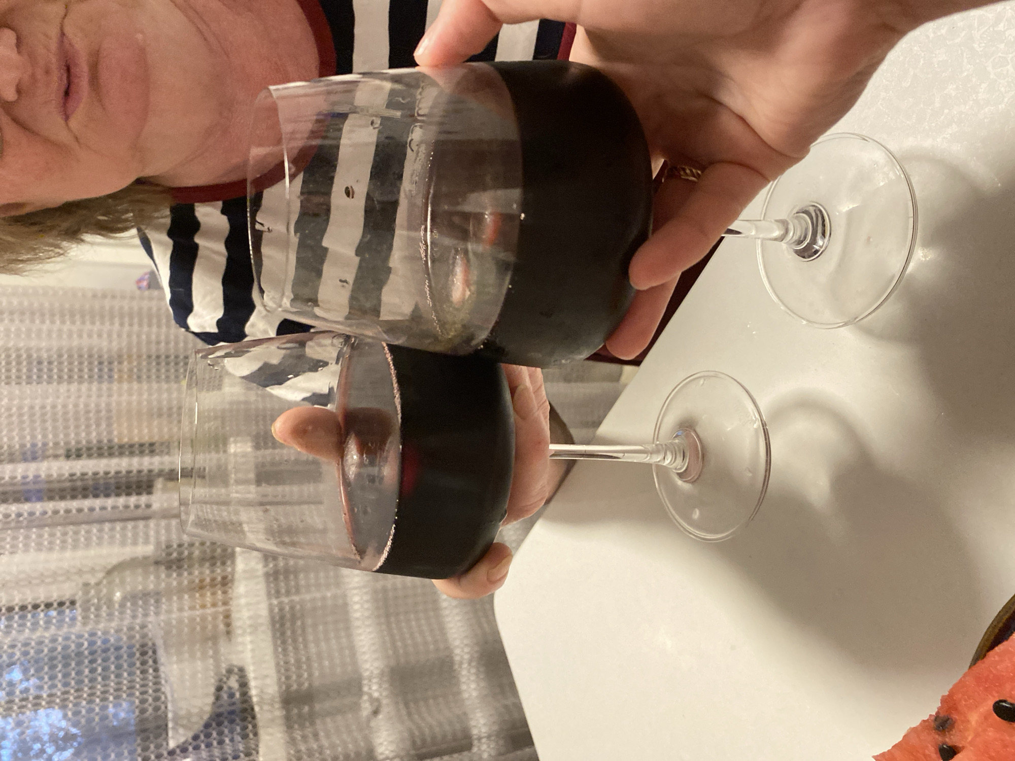 The image depicts two hands holding two wine glasses filled with red wine. A person with a striped shirt is partially visible in the background. One glass is slightly raised and closer to the camera than the other. The glasses are positioned on a white surface, with a partially visible slice of watermelon in the lower right corner. A curtain is seen in the background. The location of the image is not discernible in the city of Noviy, Russia.