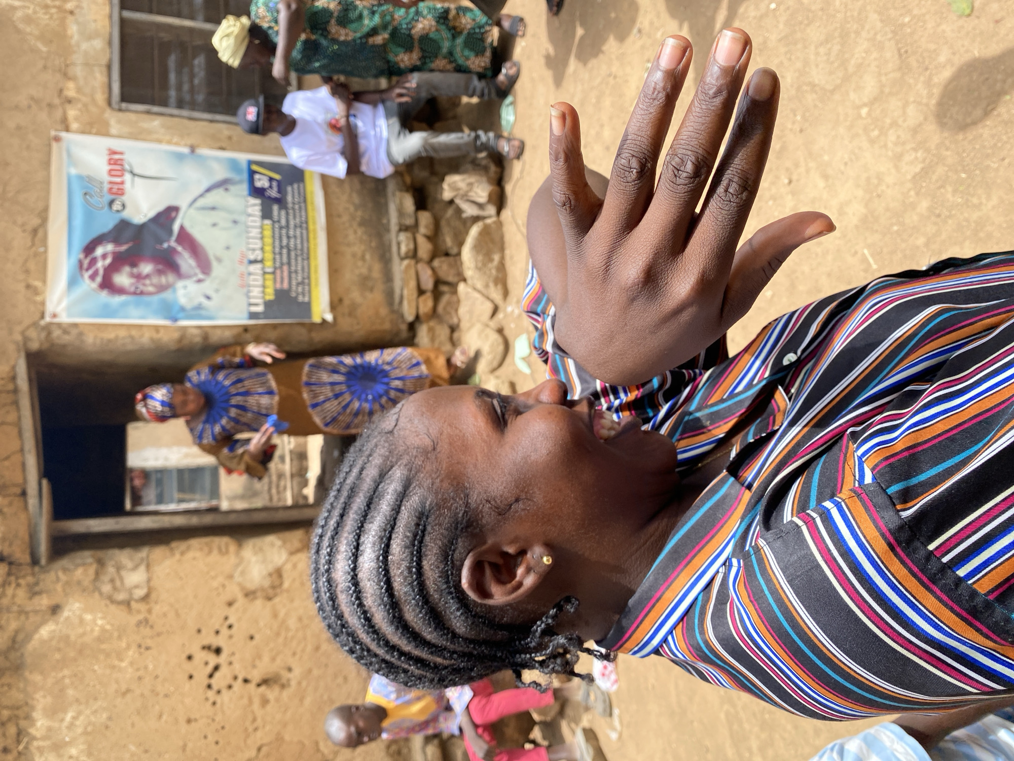 A close-up, tilted shot depicts an African woman in Vom, Nigeria, her head angled upward and to the right. She wears a striped shirt and her hair is styled in tight braids. Her right hand is raised, palm facing forward, with fingers extended and slightly apart. A gold stud earring is visible.
Behind her, a group of people are gathered near a mud-brick building. A poster on the building reads "Call to Glory" and "Linda Sunday." Additional people are partially visible, including a woman in a blue and gold patterned dress and a man wearing a white shirt and a baseball cap. The ground appears to be dirt or sand.