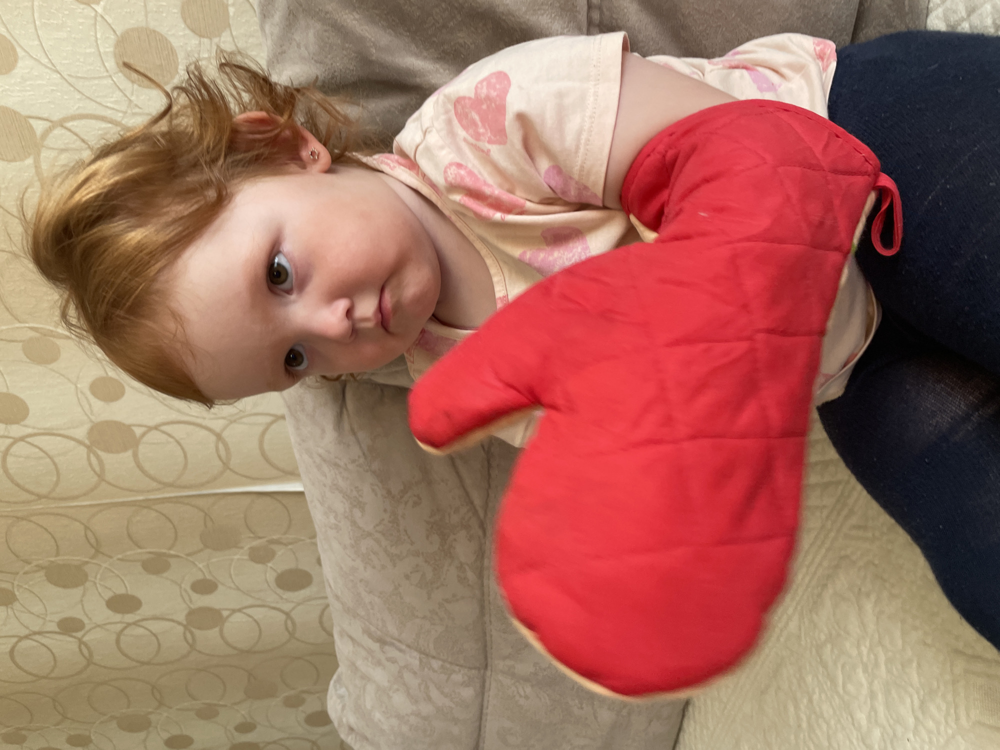 A young child with red hair is lying on a couch. The child is wearing a light pink shirt with pink hearts. The child has a red oven mitt on their right hand. The child is looking at the camera with a slight frown. The child is wearing dark blue pants. The couch is beige and has a patterned throw blanket. The location is not discernible.