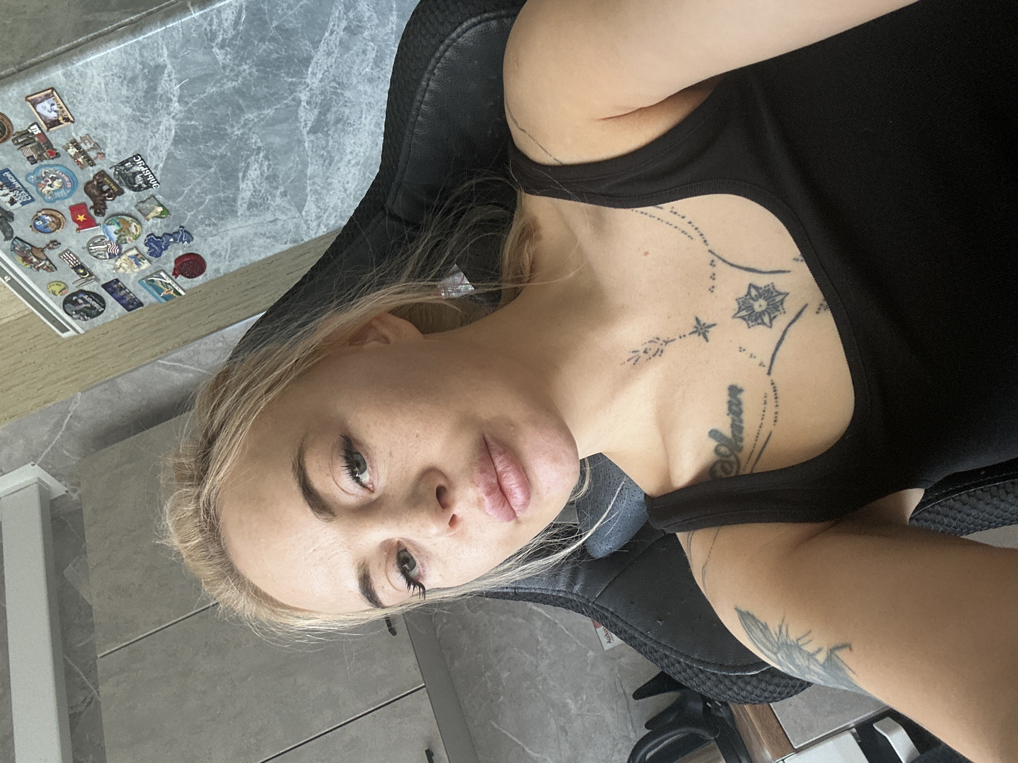 A fair-skinned woman with blonde hair and tattoos is seated in a black chair. She is wearing a black tank top. A table with numerous stickers on it is visible in the background. The precise location within the city of Высокий, Russia, cannot be determined from the provided image.