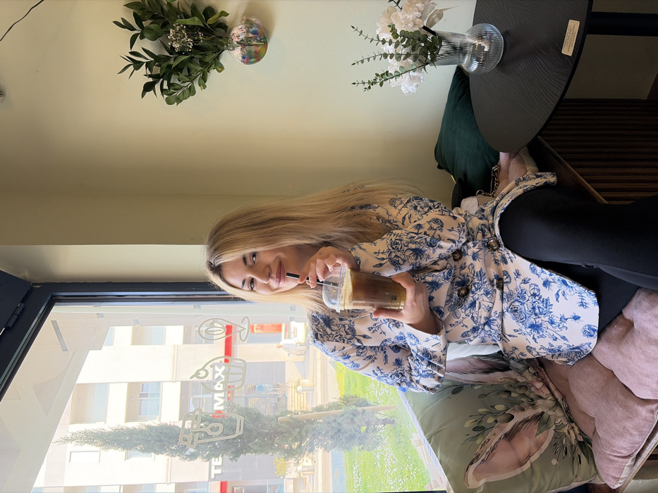 An adult female is depicted seated indoors at a cafe, positioned on a cushioned bench seat beside a large window overlooking an urban street. She has long blonde hair, styled with a center part, and is smiling directly towards the viewer. Her attire consists of a light-toned, double-breasted blazer-style top with a blue floral pattern and dark buttons, worn over black leggings. She holds a clear plastic cup containing a dark iced beverage with foam, inserting a black straw into her mouth.
The cafe interior features light green walls. The bench she occupies has a pink quilted cushion, a light green throw pillow with a rabbit motif, and a solid dark green cushion. To her right, a small, round, dark table holds a clear fluted vase containing artificial green foliage and white flowers. Wall decor includes a hanging clear glass orb with colorful contents and a cluster of green leaves with white blossoms. The large window is adorned with white vinyl decals depicting coffee cups, pastries, and location pins.
Outside the window, a tall, slender coniferous tree stands. Across the street, a multi-story building is visible, prominently displaying a red "TE MAX" sign on its ground level. This detail identifies the location as a commercial area within the city of Budva, Montenegro. The scene is illuminated by natural daylight.