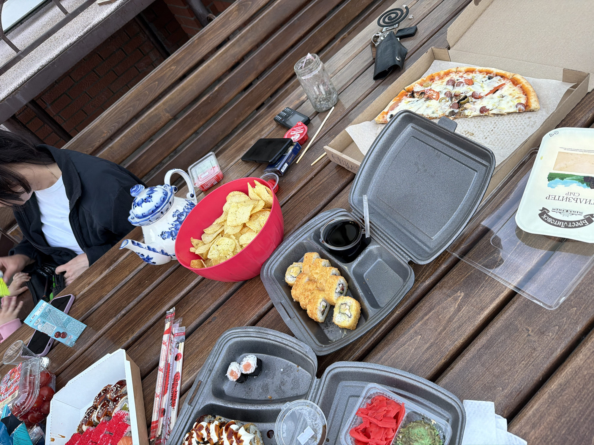 The image shows a wooden picnic table laden with food in what appears to be an outdoor setting. Dominating the scene is an assortment of food items, including sushi in various containers, a half-eaten pizza in a cardboard box, a bowl filled with potato chips, and a container of cheese. A woman wearing a black jacket and white shirt is partially visible at the left of the frame. Other notable items on the table include a blue and white teapot, a glass jar containing what appears to be plant debris, a smartphone, car keys, and various condiment containers. The picnic setting appears to be located in Chaikovsky, Russia, based on the Cyrillic text visible on the cheese container.