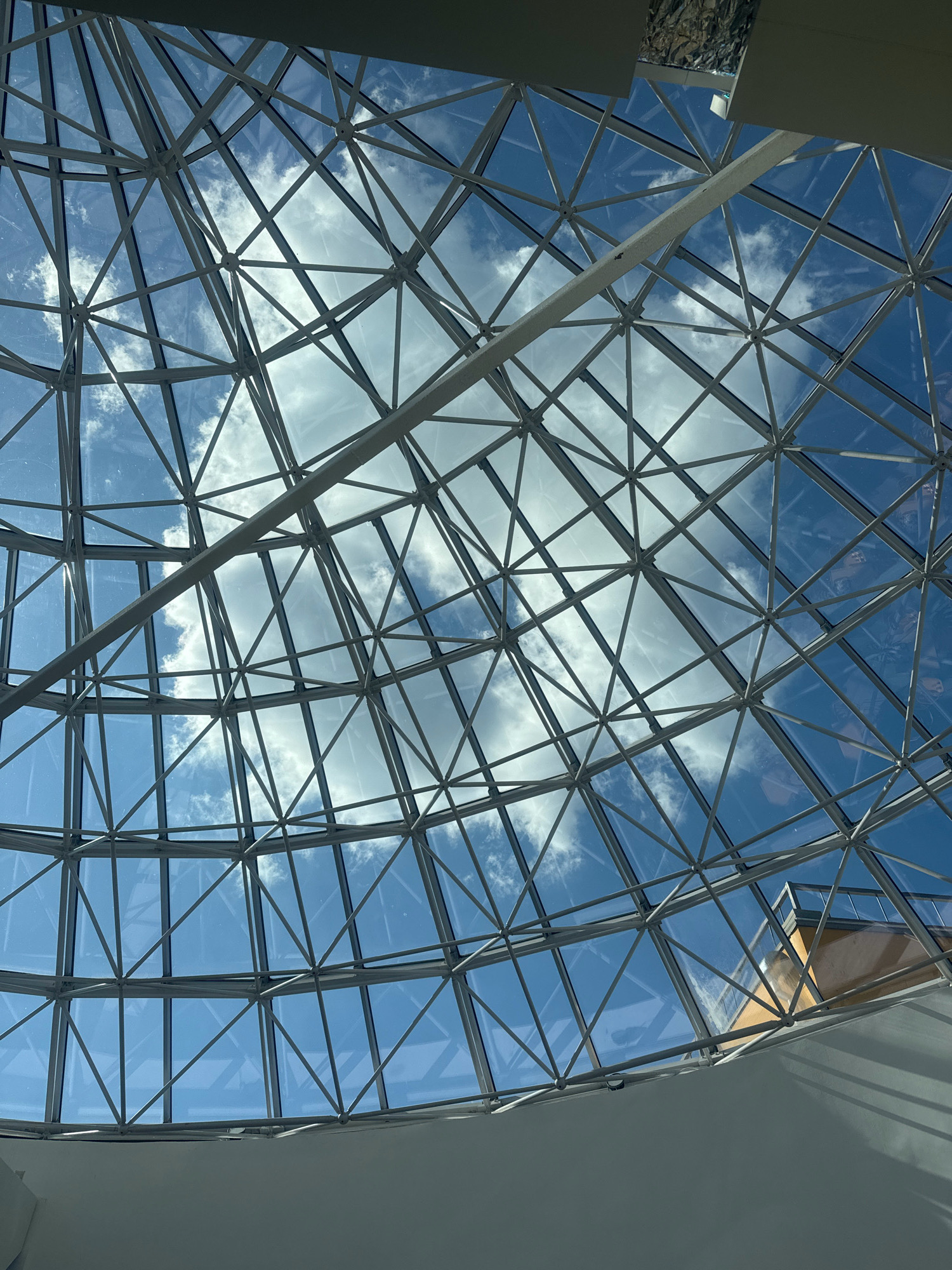 The image shows a geodesic dome ceiling made of glass and metal. The sky with scattered clouds is visible through the dome. A portion of a building with an orange facade is visible in the upper right corner. The image is taken from an interior space looking upwards at the dome. There is no information in the image that would indicate its location in Irkutsk, Russia.