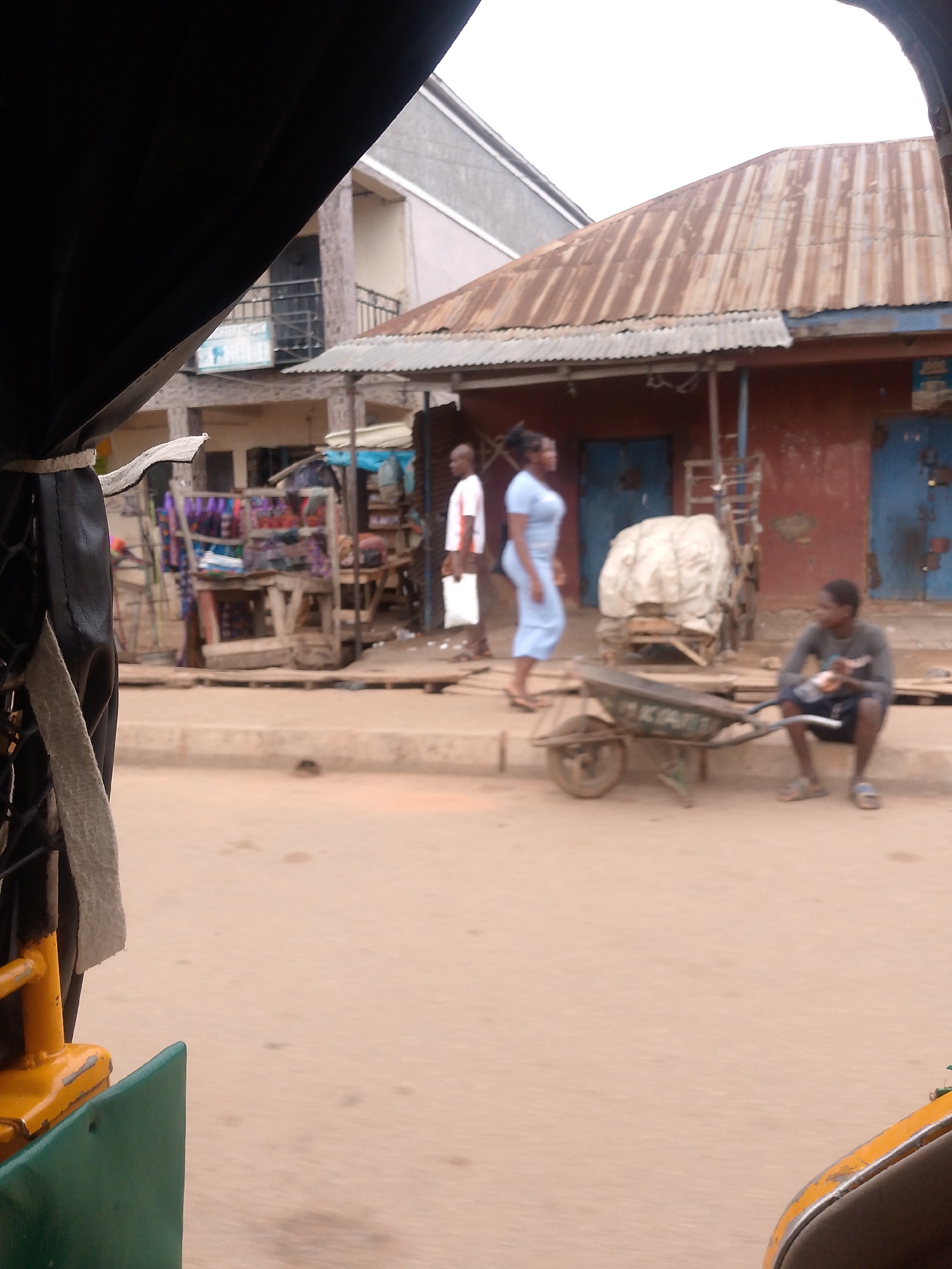 The image depicts a street scene in Bukuru, Nigeria, taken from inside a motorized tricycle (keke). In the background, there is a building with a corrugated metal roof and two closed blue doors. In front of it, a man in a white shirt stands with a white bag. A woman in a blue dress stands next to him. A man sits on the curb next to a wheelbarrow, appearing to be eating. In the foreground, a portion of the keke's black canvas roof, yellow frame, and green side panel are visible. Across the street are market stalls selling colorful textiles and other items.